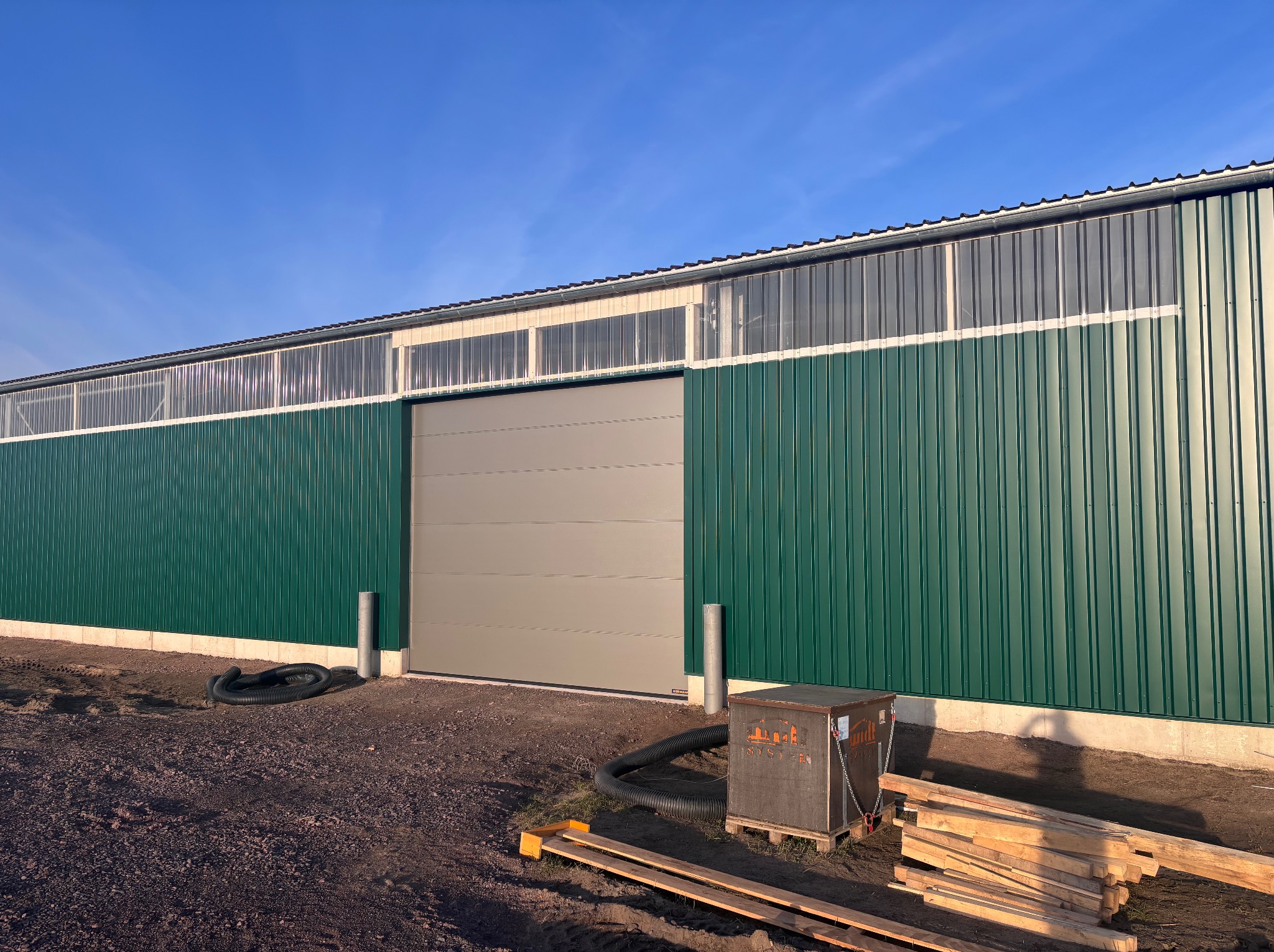 Zielona hala z szarą bramą przemysłową segmentową, widoczne elementy konstrukcyjne i materiały budowlane na ziemi. Błękitne niebo w tle.