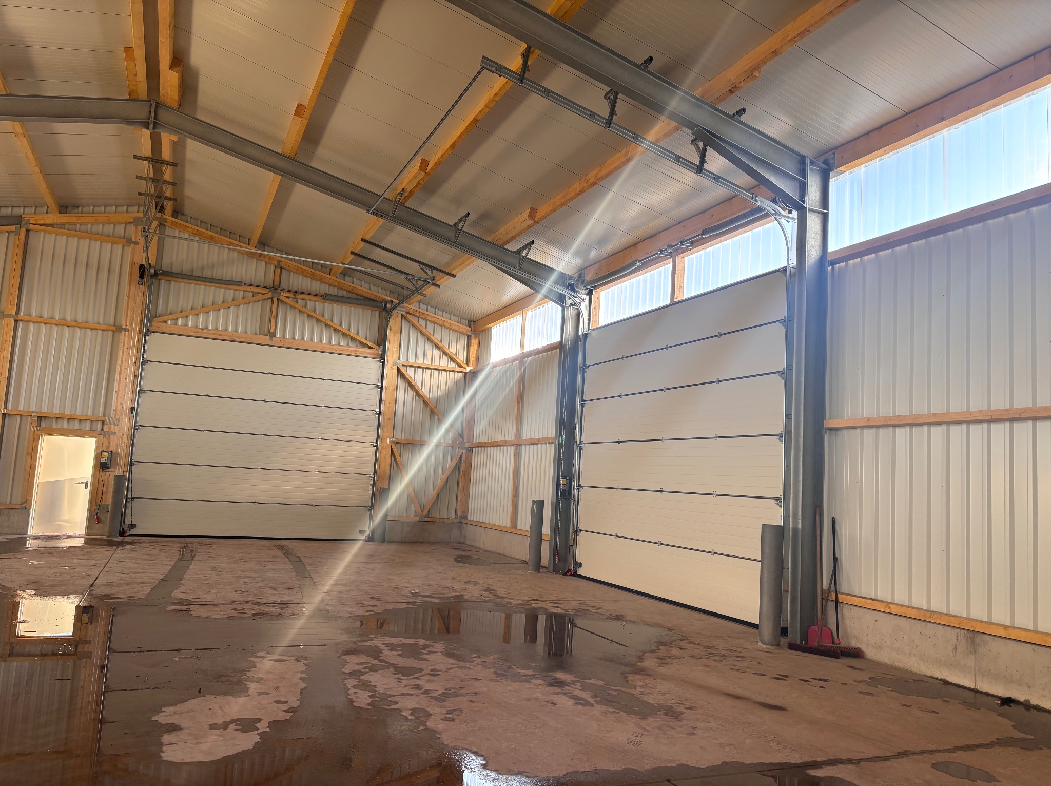 Wnętrze hali z zamontowanymi segmentowymi bramami przemysłowymi w kolorze białym. Widoczne drewniane elementy konstrukcyjne i wpadające światło słoneczne.