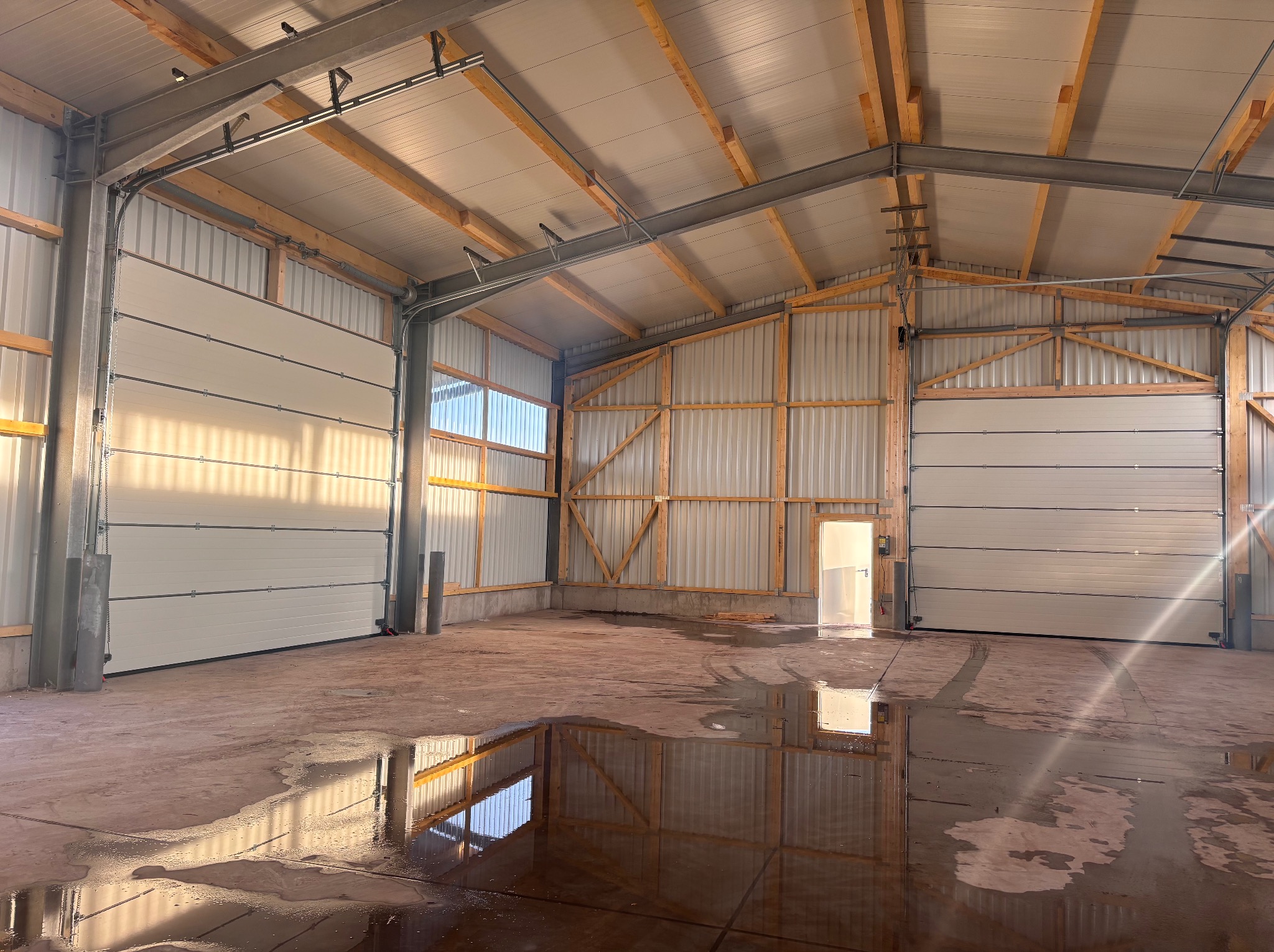 Wnętrze hali z zamontowanymi segmentowymi bramami przemysłowymi, widoczne drewniane elementy konstrukcyjne i mokra posadzka z odbiciami.