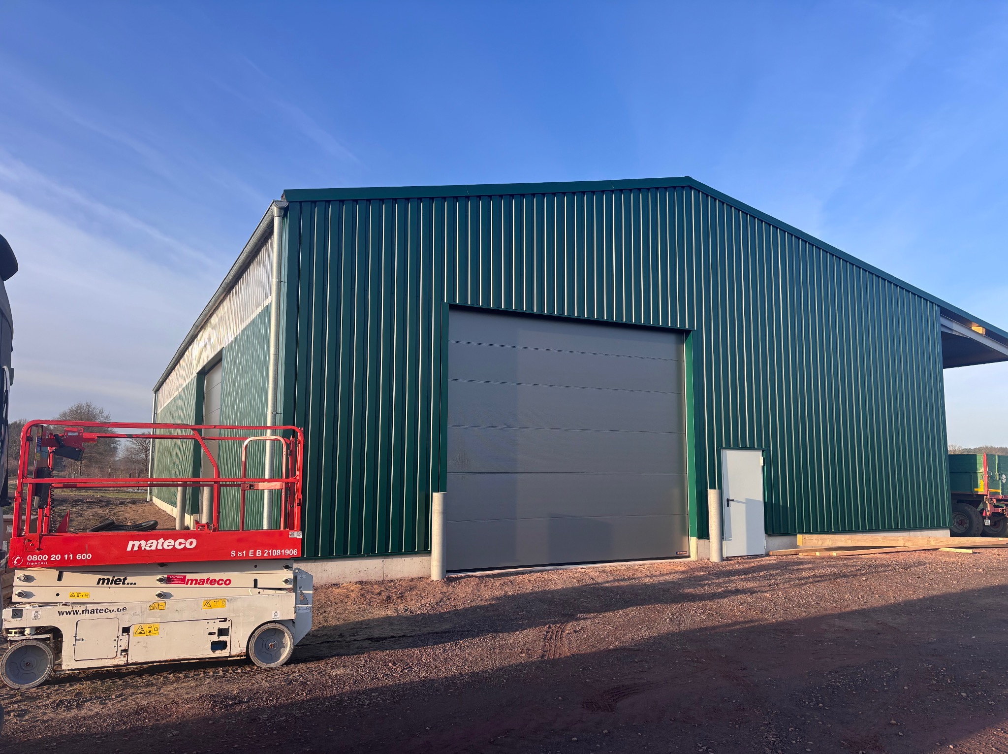 Zielony budynek przemysłowy z szarą bramą segmentową, widoczny podnośnik koszowy Mateco. Nowoczesna hala z blachy trapezowej w słoneczny dzień.