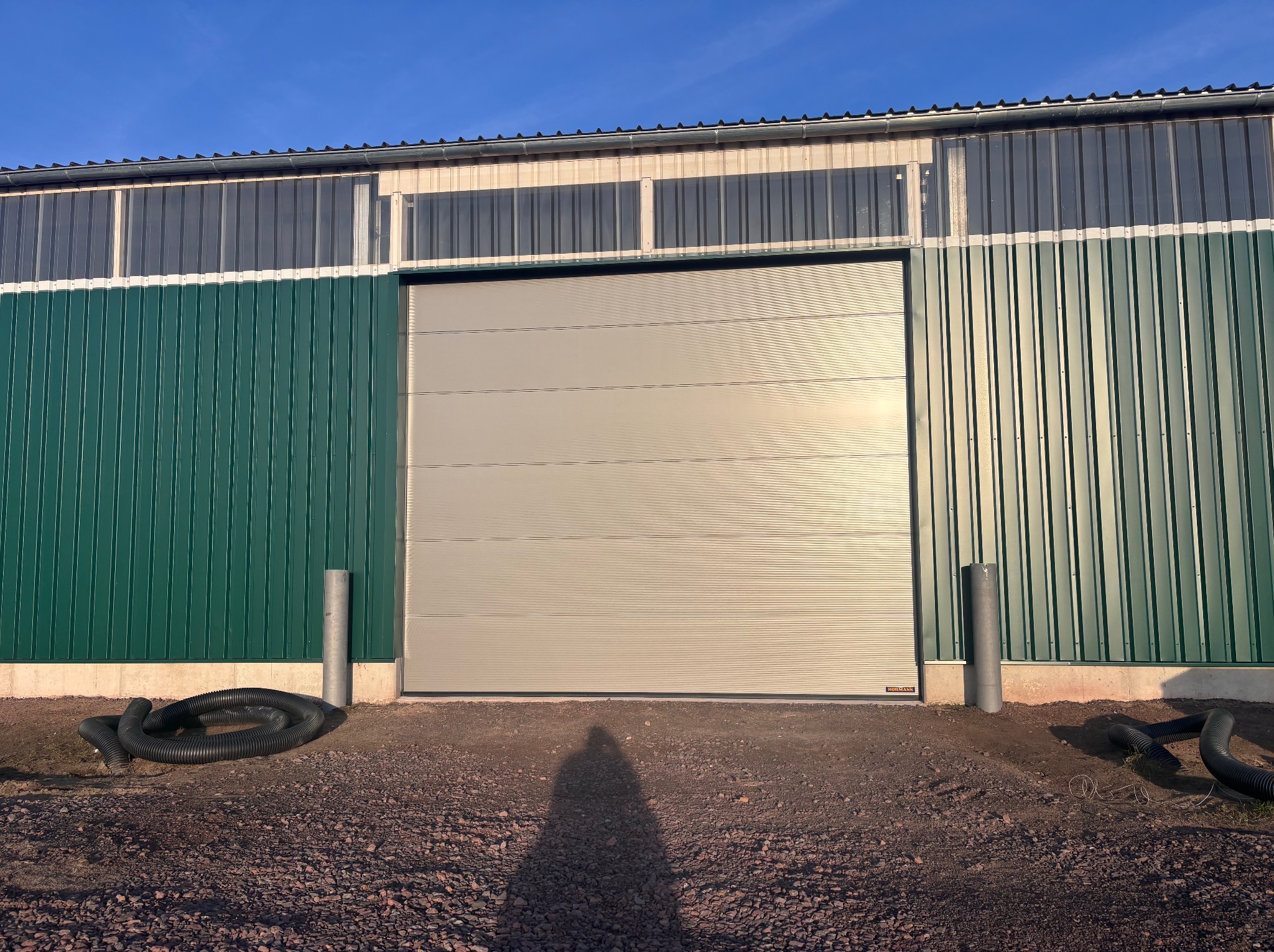 Szara brama przemysłowa segmentowa w zielonej hali z blachy trapezowej, widoczne rury drenażowe i cień osoby na żwirowym podłożu. Brama marki Hormann.