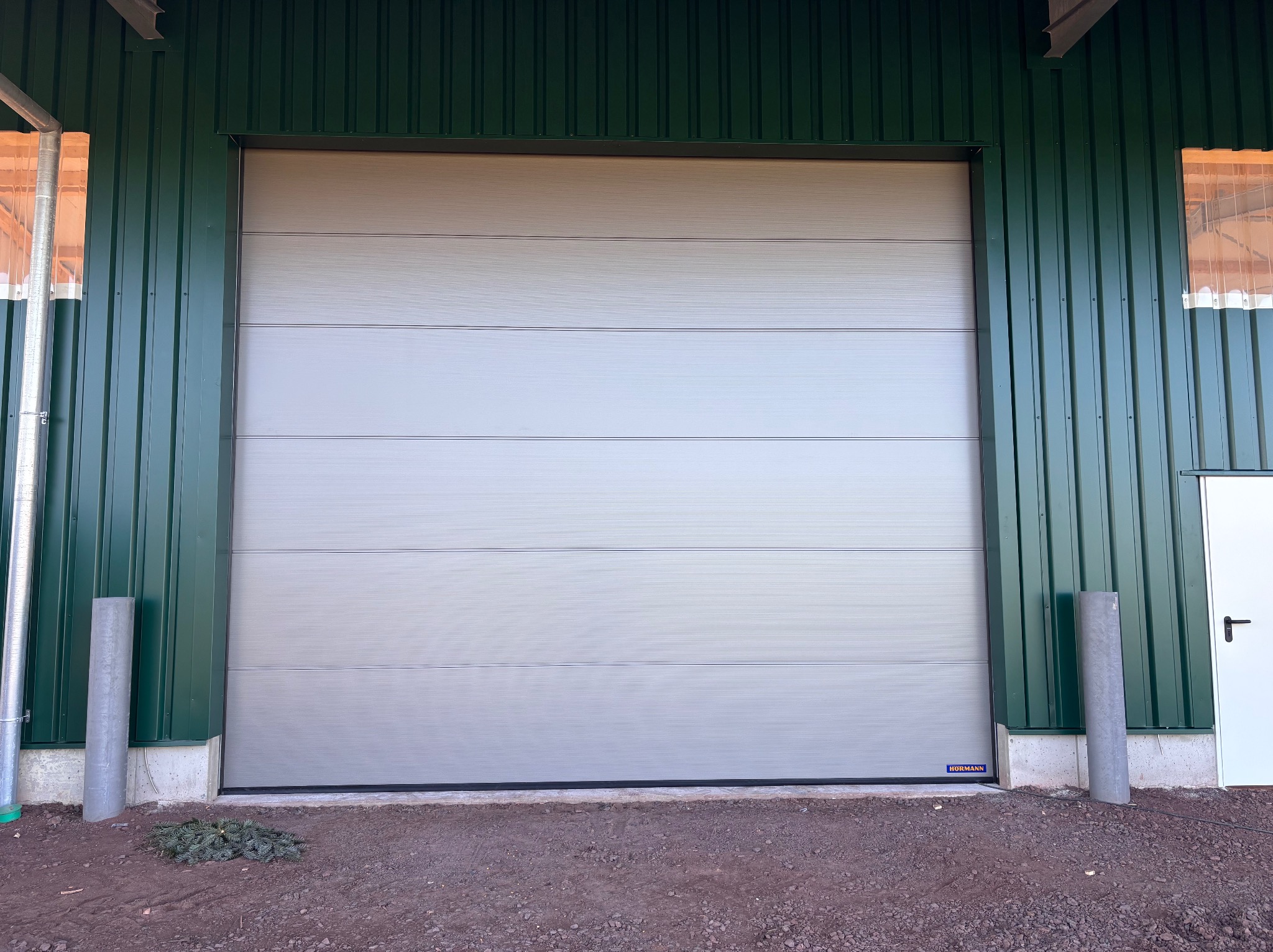 Zamknięta, segmentowa brama garażowa w kolorze jasnoszarym, z widocznym logo producenta, zamontowana w zielonej hali z blachy trapezowej. U dołu widoczny fragment ziemi.