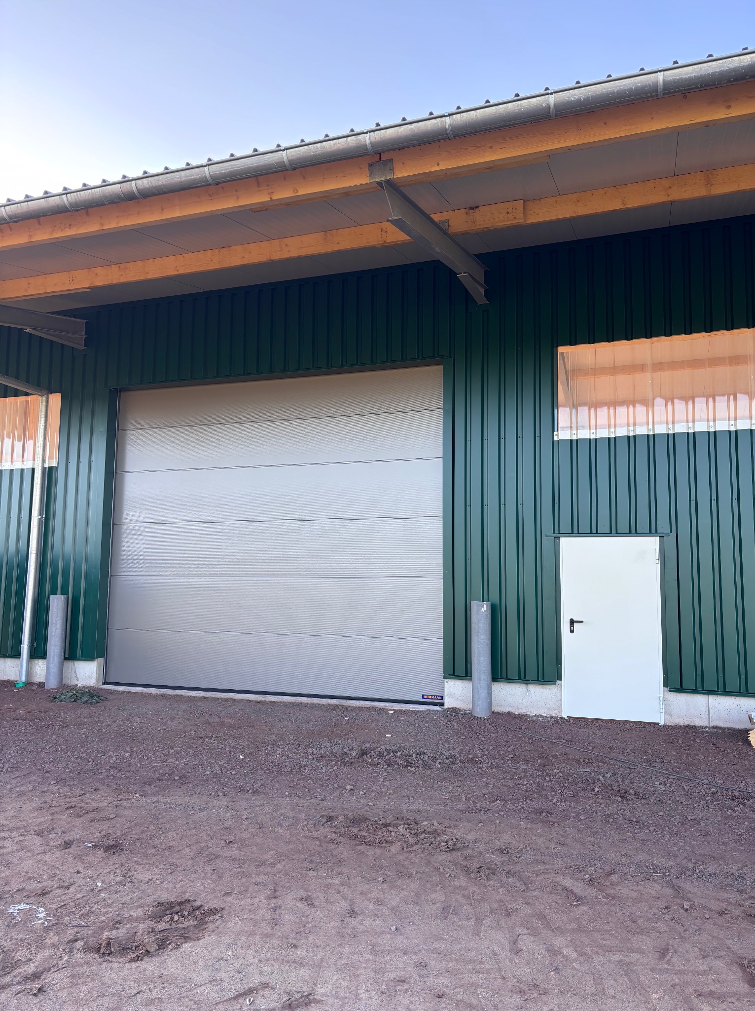 Szara brama segmentowa w zielonej hali przemysłowej z dodatkowym białym wejściem. Widoczne elementy konstrukcyjne dachu i podłoże z ziemi.