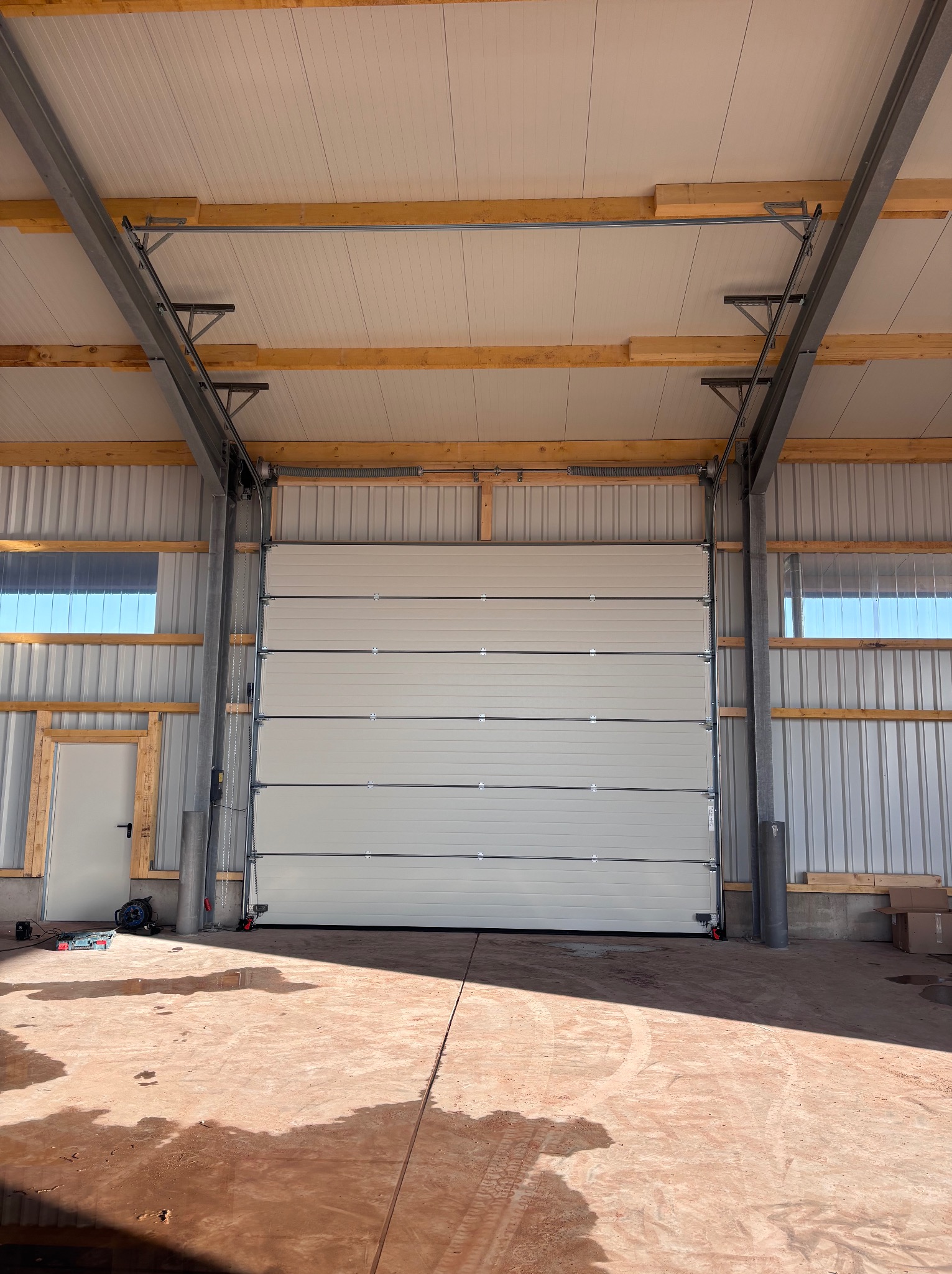 Zamknięta, segmentowa brama garażowa wewnątrz hali z widoczną konstrukcją stalową i drewnianą. Na podłodze widoczne ślady po budowie. Brama w kolorze jasnym.