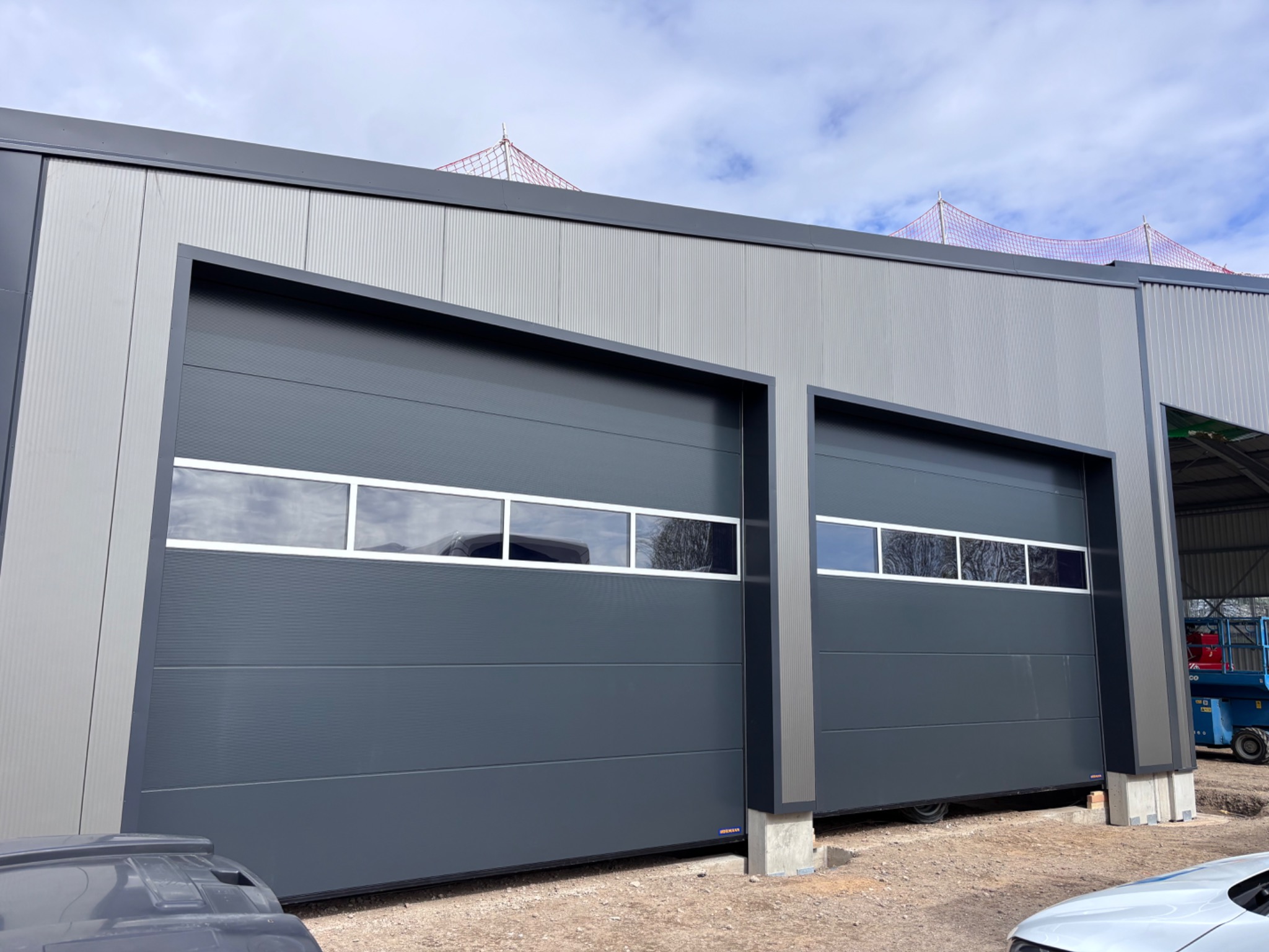 Dwie szare bramy garażowe segmentowe z oknami, zamontowane w szarym budynku przemysłowym. Widoczna konstrukcja budynku i fragmenty pojazdów.