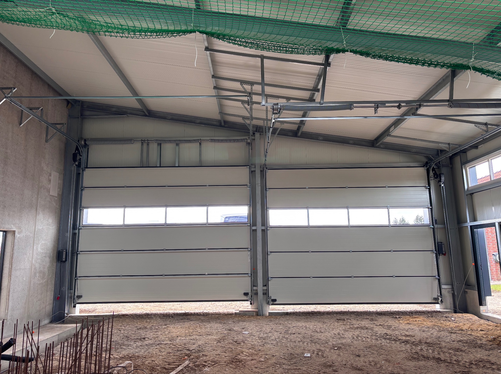 Dwie segmentowe bramy przemysłowe z oknami, częściowo otwarte, w surowym wnętrzu hali z widoczną konstrukcją dachu i elementami instalacji. Widoczny piasek na podłodze.