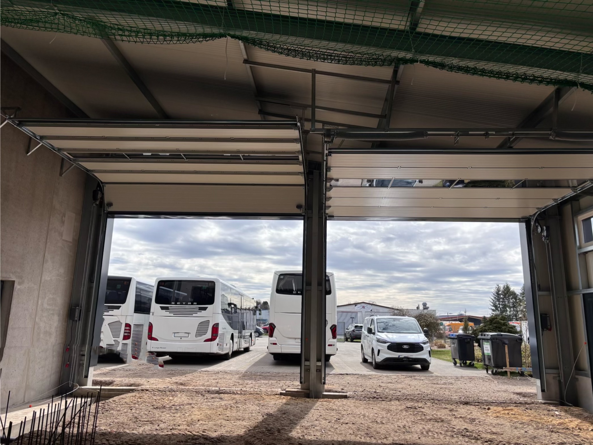 Otwarta brama przemysłowa z widokiem na autobusy i samochód osobowy na zewnątrz. Widoczna konstrukcja bramy i fragment dachu z siatką zabezpieczającą.