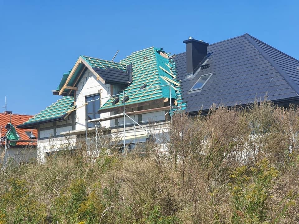 Dom w trakcie krycia dachu: widoczne rusztowania, konstrukcja dachu z zieloną folią i fragment gotowego dachu z czarną dachówką. Tło: błękitne niebo.