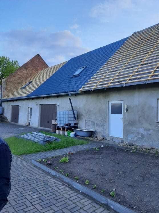 Częściowo pokryty, niebieski dach z oknem dachowym na budynku. Widoczne drewniane łaty na niezakończonej części dachu. Fragment muru z cegły.