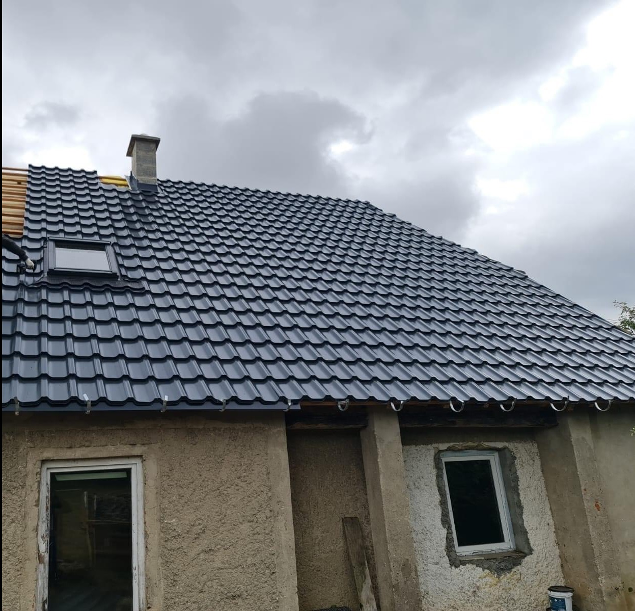 Ciemny dach z dachówką ceramiczną, widok z dołu na dom z oknami i kominem. Częściowo widoczne drewniane elementy konstrukcji dachu.