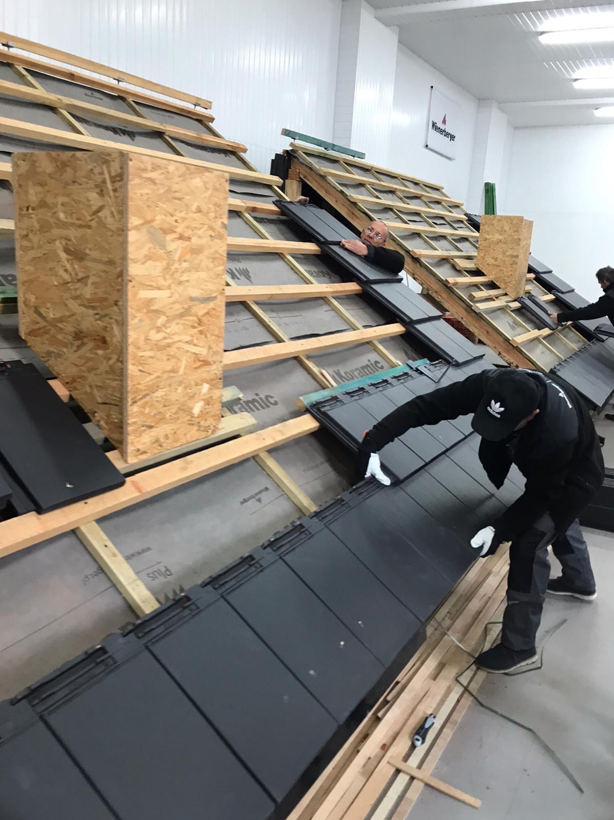 Montaż czarnej dachówki ceramicznej na drewnianej konstrukcji dachu przez dekarzy w białych rękawicach. Widoczny komin i logo producenta.