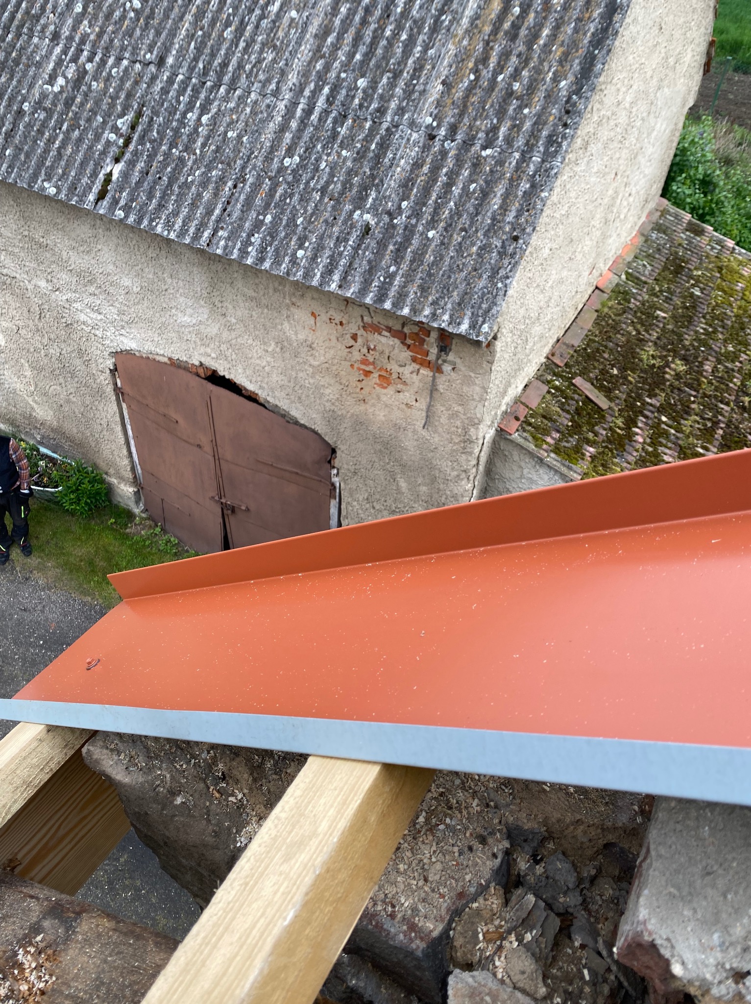 Montaż nowej, pomarańczowej obróbki blacharskiej dachu na tle starego budynku z eternitem i ceglanymi elementami. Widoczne drewniane elementy konstrukcji.