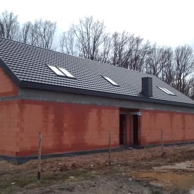 Budynek w trakcie budowy z szarym dachem i oknami dachowymi. Widoczne czerwone ściany z pustaków i komin. W tle drzewa bez liści.