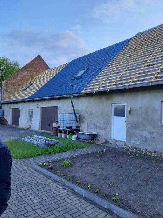 Częściowo pokryty, niebieski dach z oknem dachowym na budynku gospodarczym. Widoczne krokwie i stara część dachu z cegły. Na podwórku zbiornik na wodę i drabina.