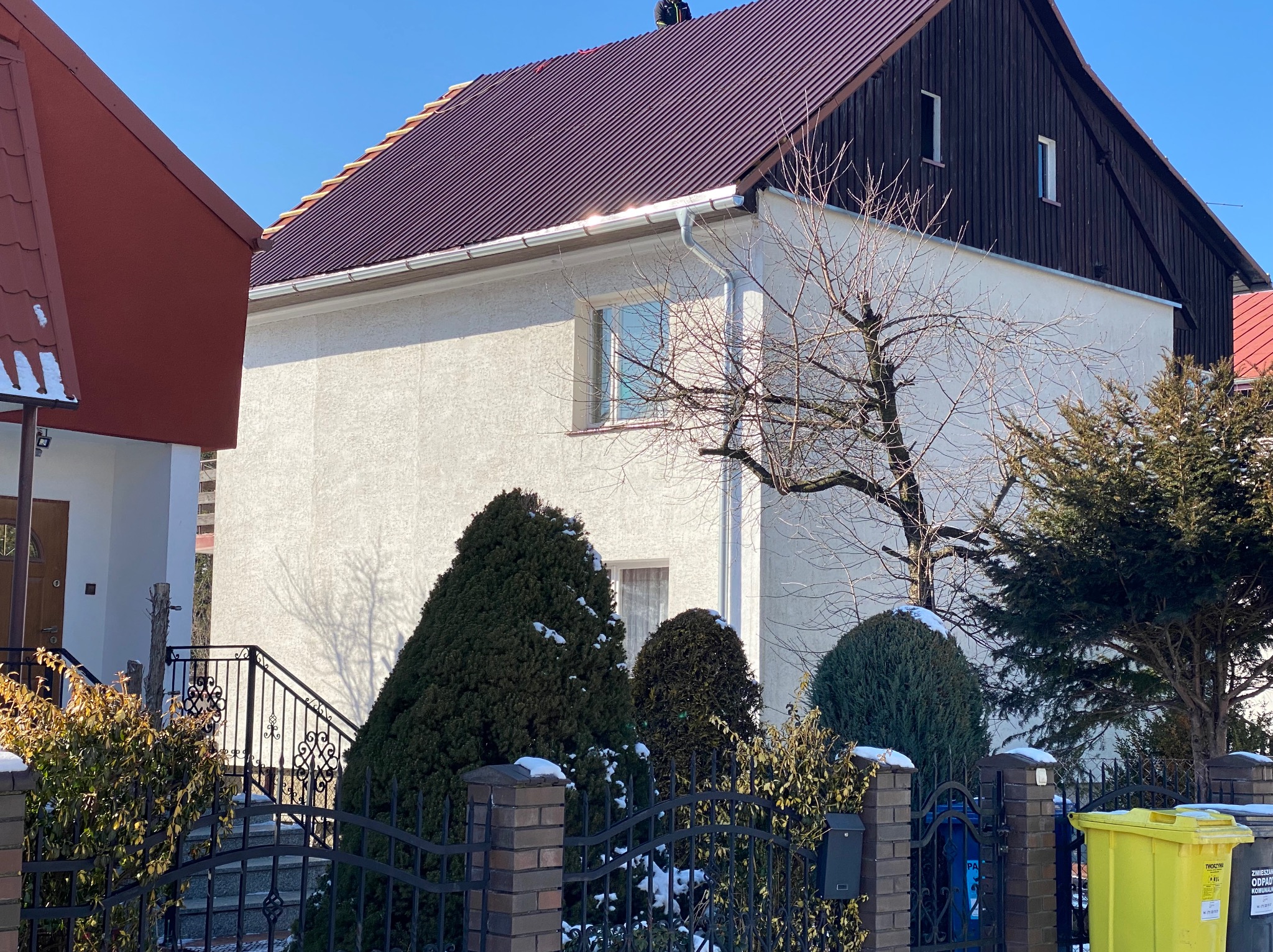 Dwupiętrowy dom z ciemnoczerwonym dachem i białymi ścianami, widoczny zza ogrodzenia z ceglanymi słupkami i zielenią. Rynny w kolorze srebrnym.