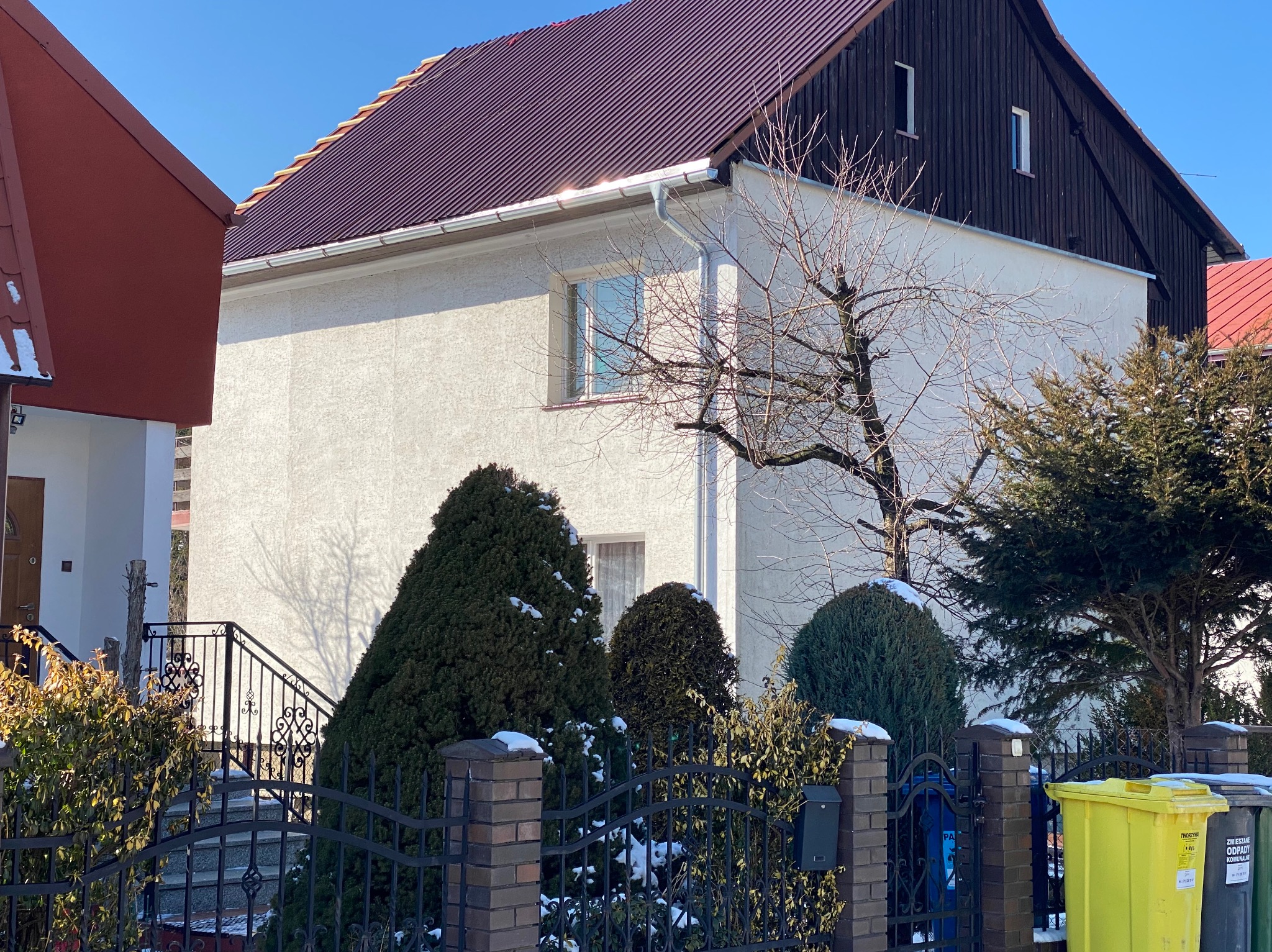 Dom jednorodzinny z ciemnobrązowym dachem z blachy trapezowej i białymi rynnami. Widoczne krzewy iglaste i drzewo bez liści. Częściowo widoczne ogrodzenie z metaloplastyki.