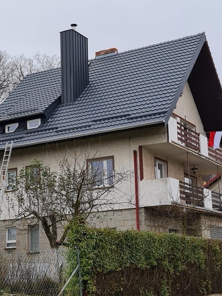 Dom jednorodzinny z nowym, ciemnoszarym dachem z blachy trapezowej i kominem. Na dachu widoczna drabina. Tło: pochmurne niebo i drzewa bez liści.