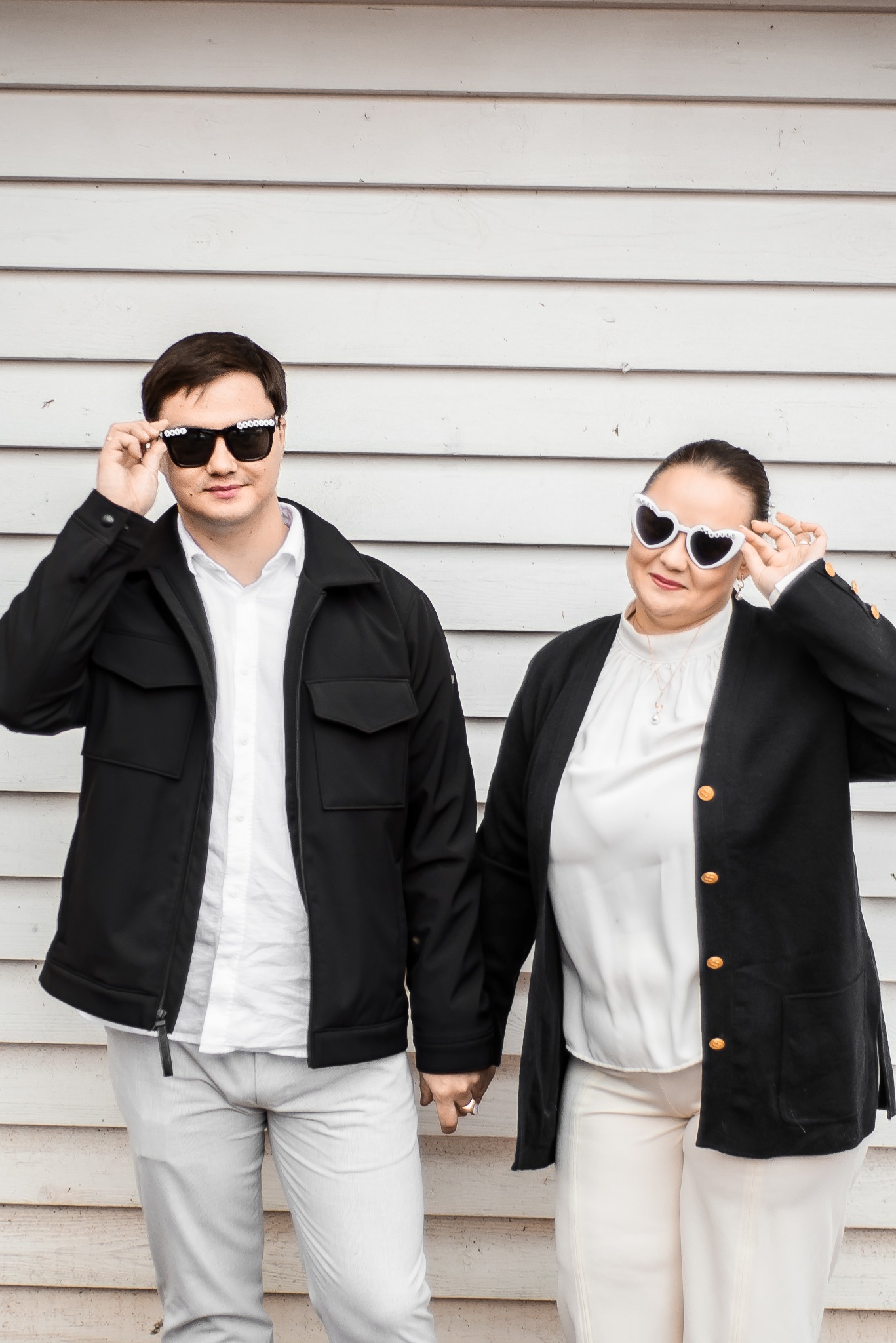 Para trzymająca się za ręce, oboje w okularach przeciwsłonecznych z ozdobami, na tle białej, drewnianej ściany. Uśmiechnięci, w eleganckich strojach.