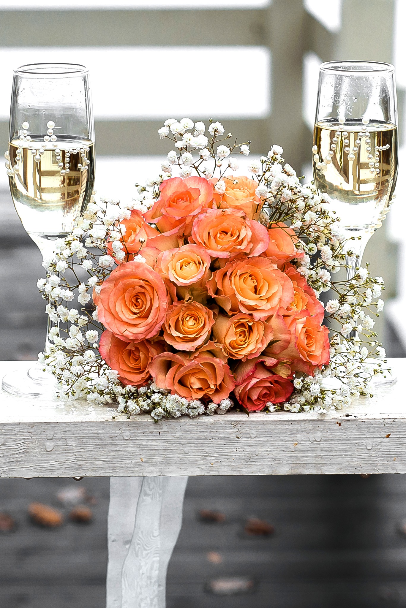 Dwa kieliszki szampana z perłami i bukiet pomarańczowych róż z gipsówką na białym tle. Romantyczna kompozycja ślubna, delikatne światło.