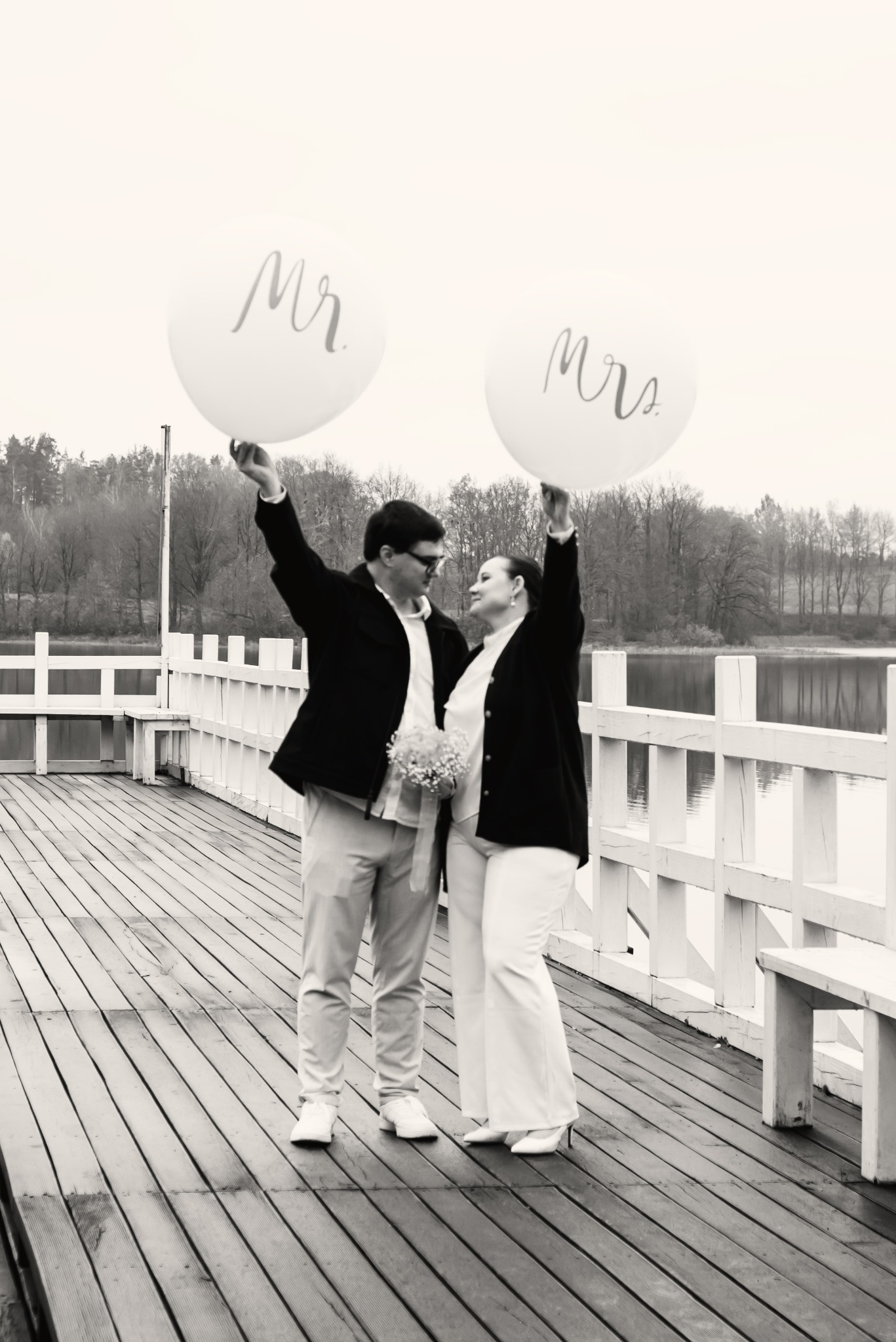 Czarno-białe zdjęcie pary młodej na drewnianym pomoście z balonami 'Mr' i 'Mrs' w rękach, z widokiem na jezioro i las w tle. Romantyczna atmosfera.