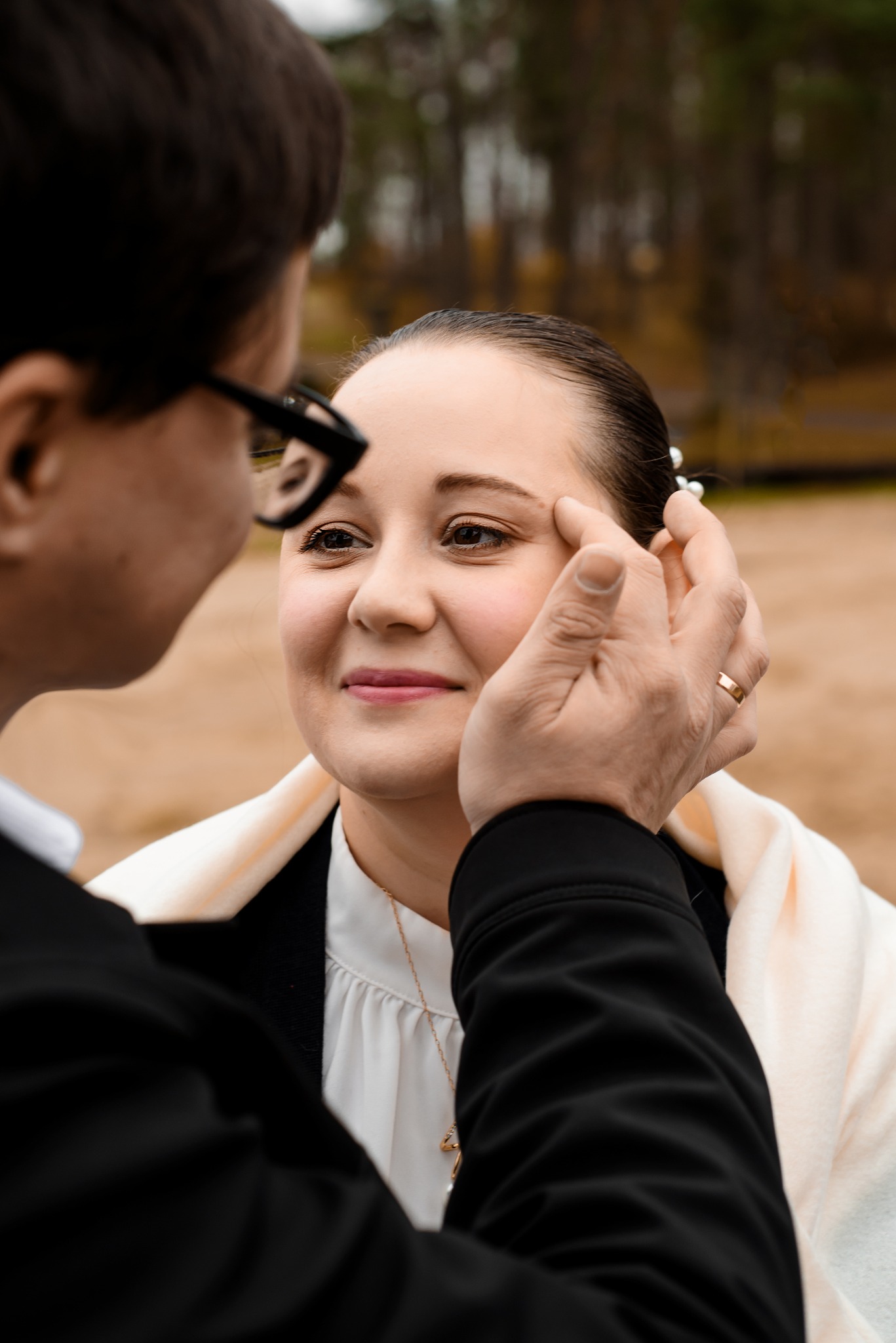 Uśmiechnięta panna młoda z delikatnym makijażem, dotykana przez pana młodego z obrączką na palcu, w plenerze z rozmytym tłem leśnym.