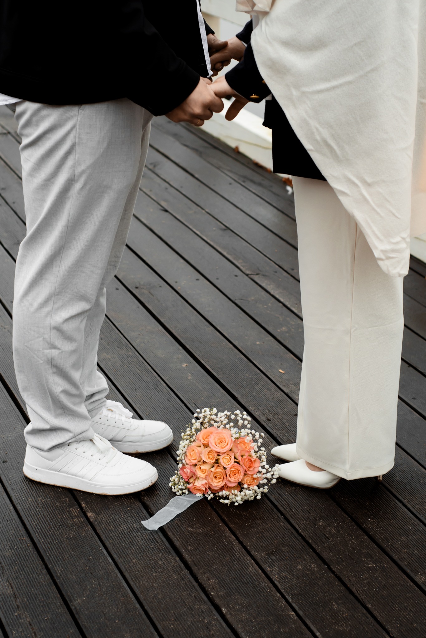 Para młoda trzymająca się za ręce, bukiet pomarańczowych róż leży na drewnianym pomoście. Ujęcie od dołu, widoczne nogi i buty pary.