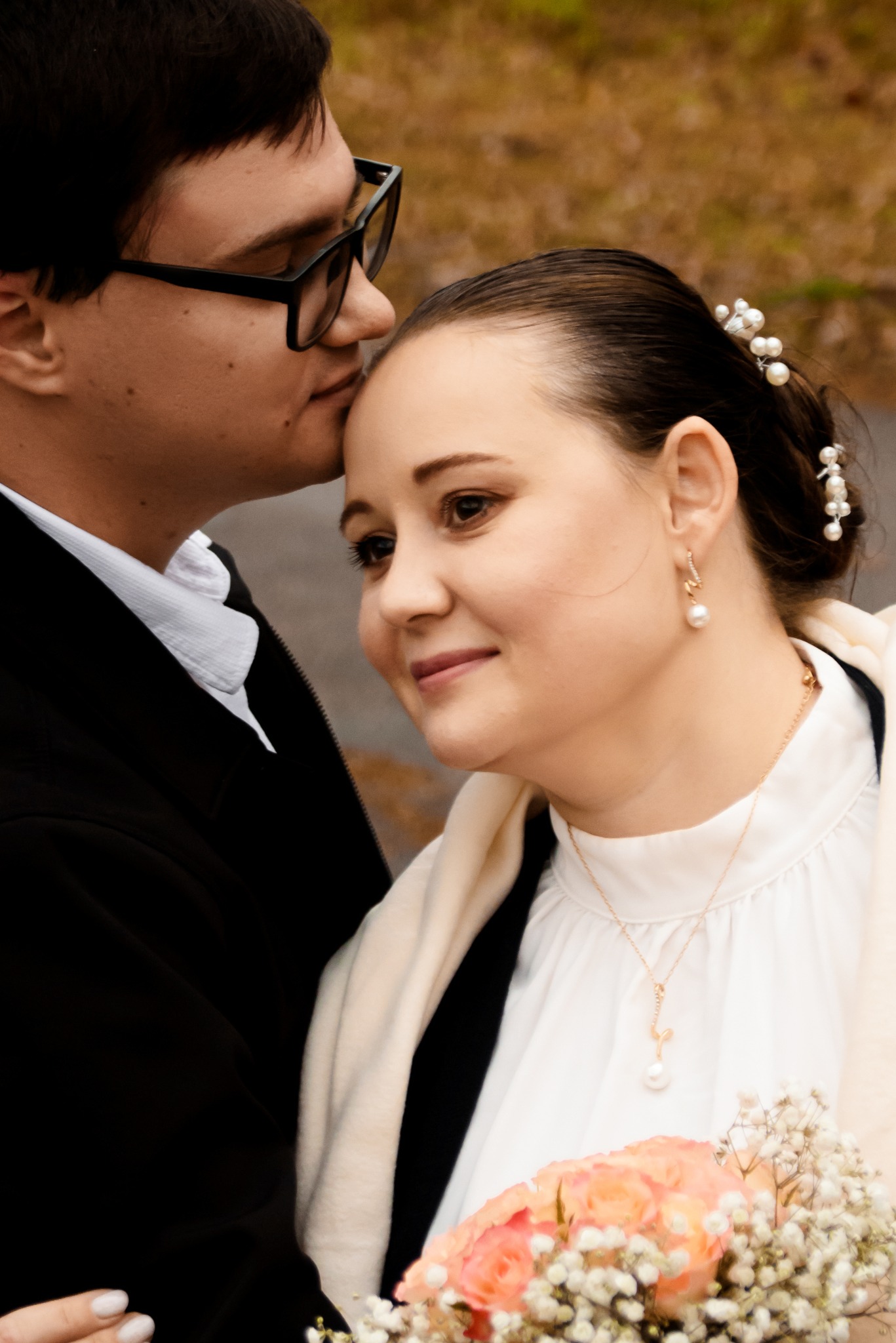 Uśmiechnięta panna młoda z bukietem, w objęciach pana młodego w okularach, który całuje ją w głowę. Delikatna biżuteria i kwiaty we włosach.