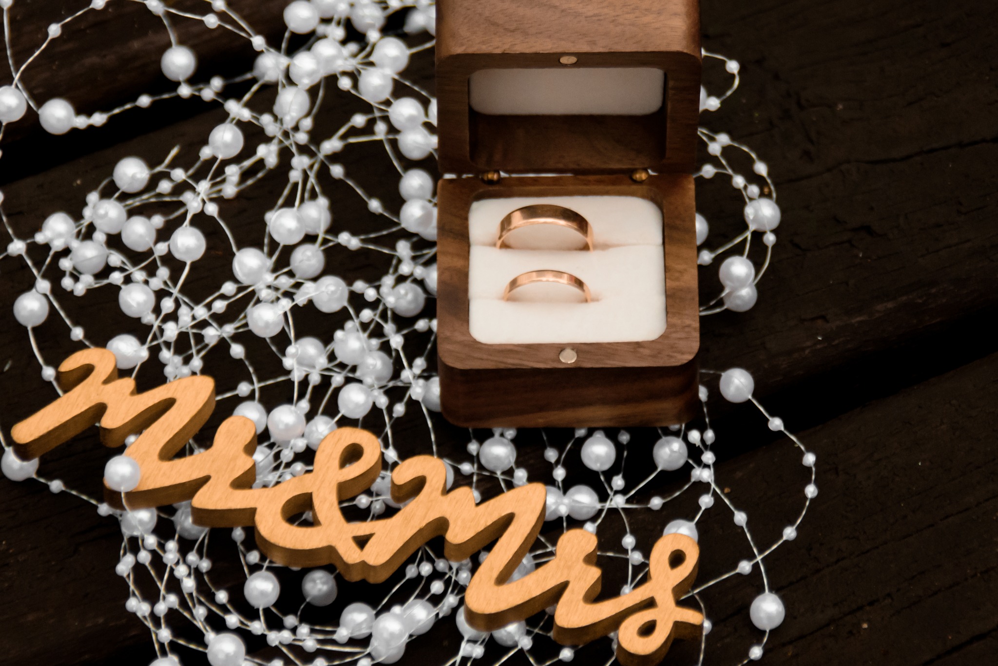 Otwarta drewniana szkatułka z obrączkami na białym tle z pereł i napisem 'Mr & Mrs' na ciemnym drewnianym stole. Ujęcie z góry, ciepłe światło.