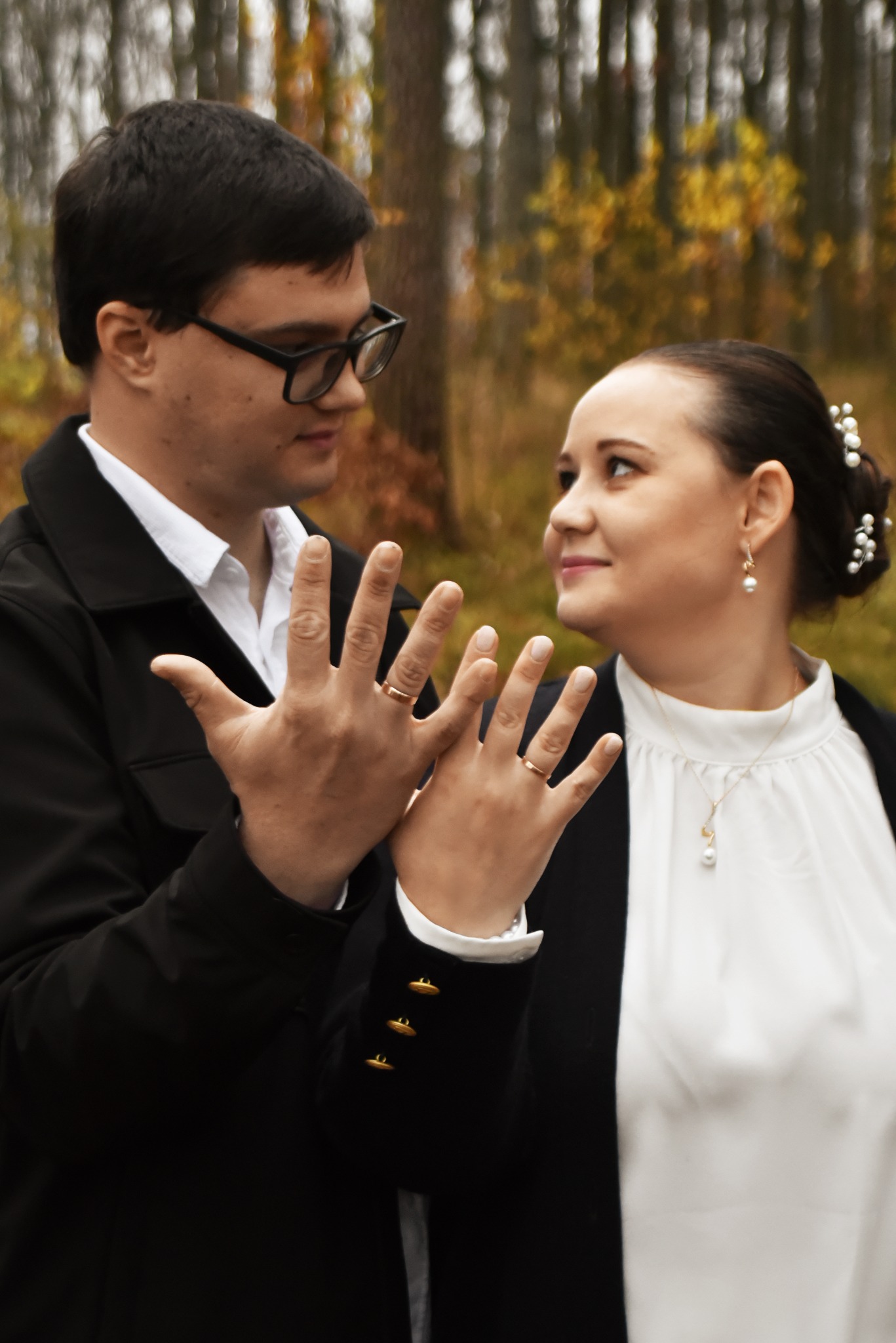 Uśmiechnięta para w jesiennej scenerii prezentuje dłonie z obrączkami, symbolizując zawarcie związku małżeńskiego. Delikatne światło podkreśla detale biżuterii.