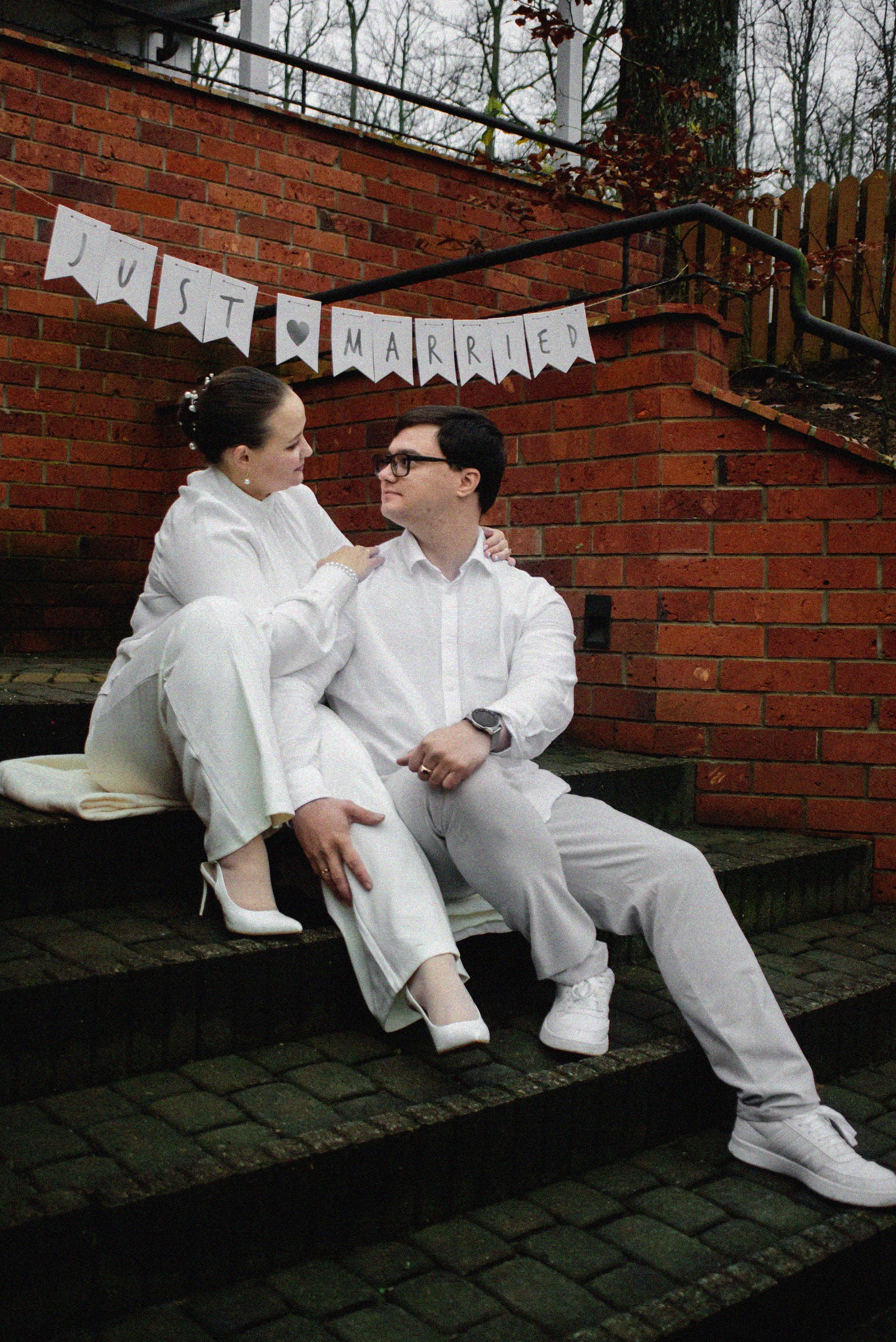 Uśmiechnięta para młoda w bieli siedzi na schodach z ceglanym murem w tle, nad nimi baner 'Just Married'. Ona w szpilkach, on w sportowych butach. Romantyczny kadr.