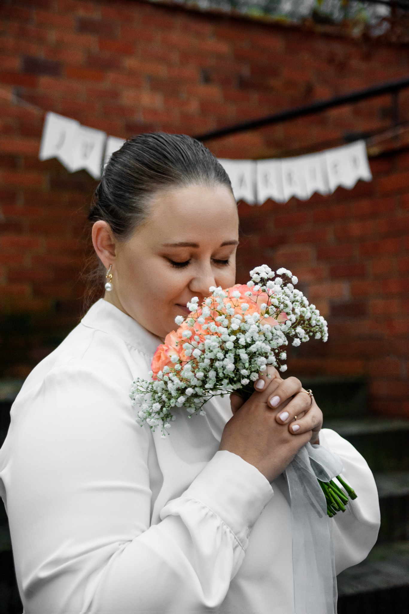 Panna młoda w białej bluzce z bukietem różowych róż i gipsówki na tle ceglanej ściany z dekoracyjnym banerem. Delikatny makijaż i perłowe kolczyki.