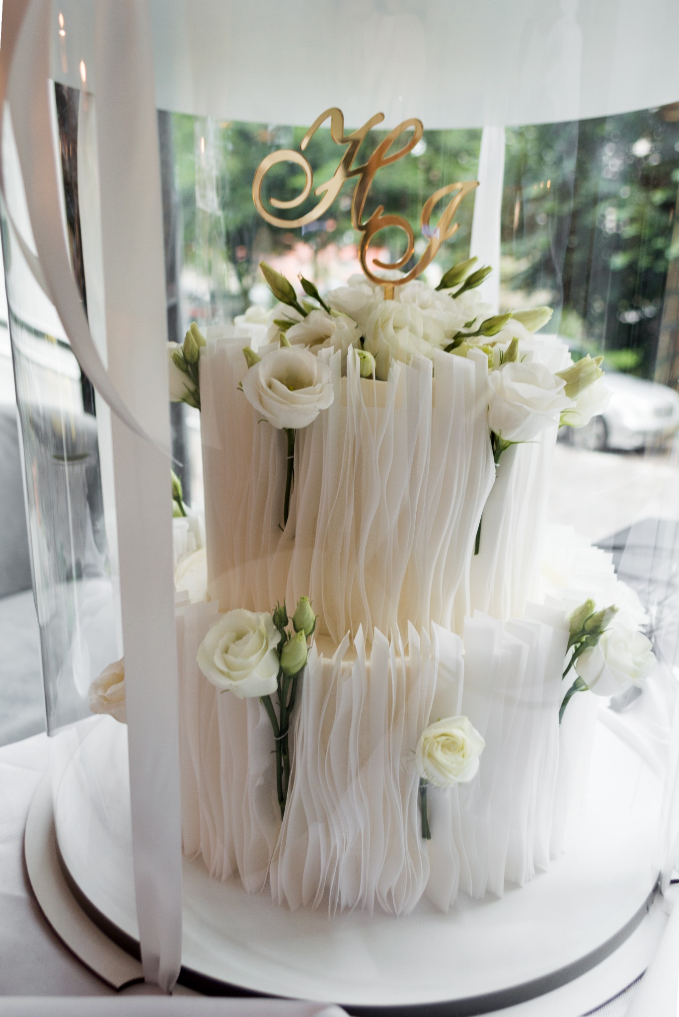 Dwupoziomowy tort weselny w białej kolorystyce, zdobiony żywymi kwiatami i topperem z inicjałami, umieszczony w przezroczystej osłonie. Delikatne światło dzienne.