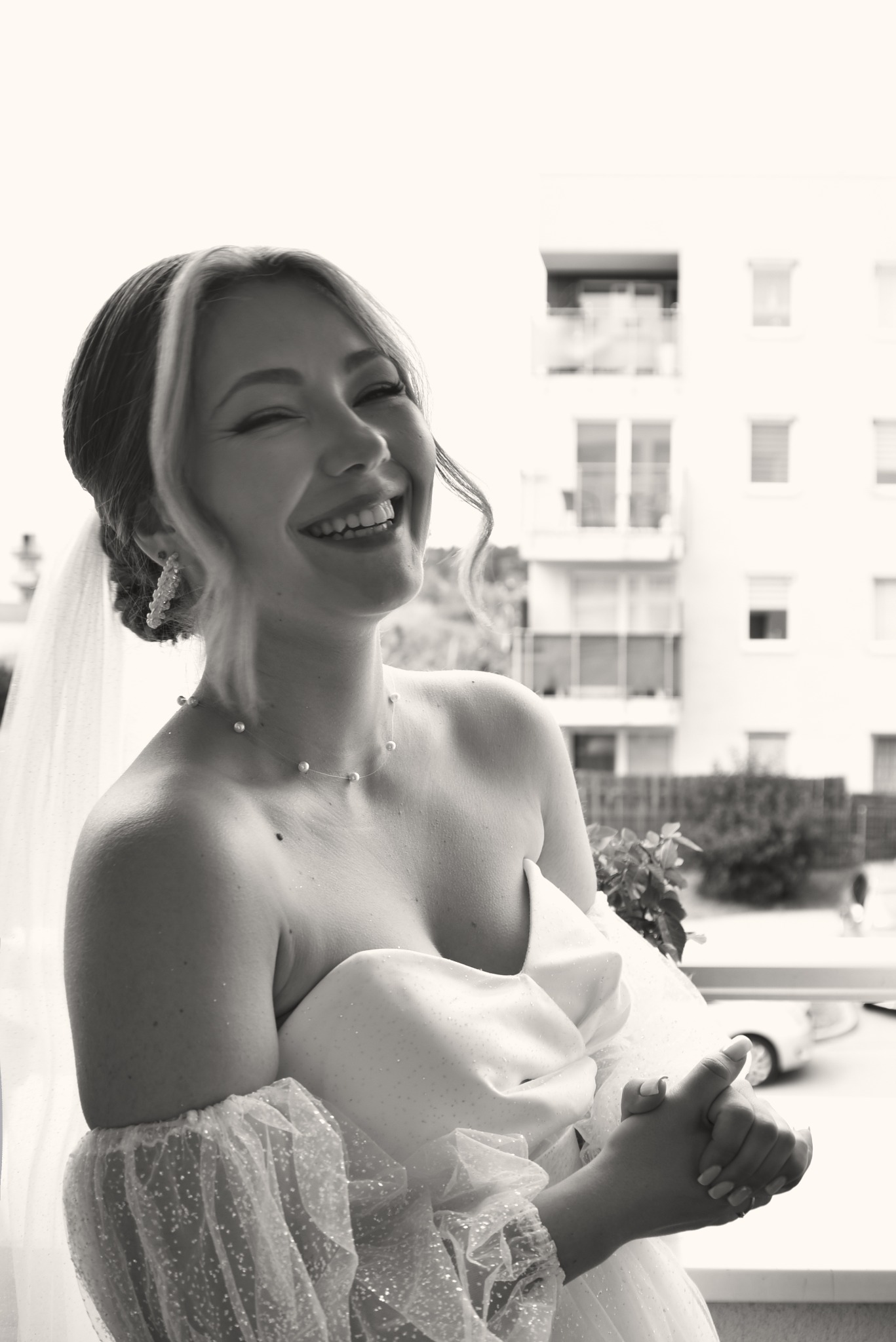 Uśmiechnięta panna młoda w sukni bez ramiączek, z perłowym naszyjnikiem i welonem, na tle budynku. Czarno-biała fotografia ślubna z subtelnym oświetleniem.