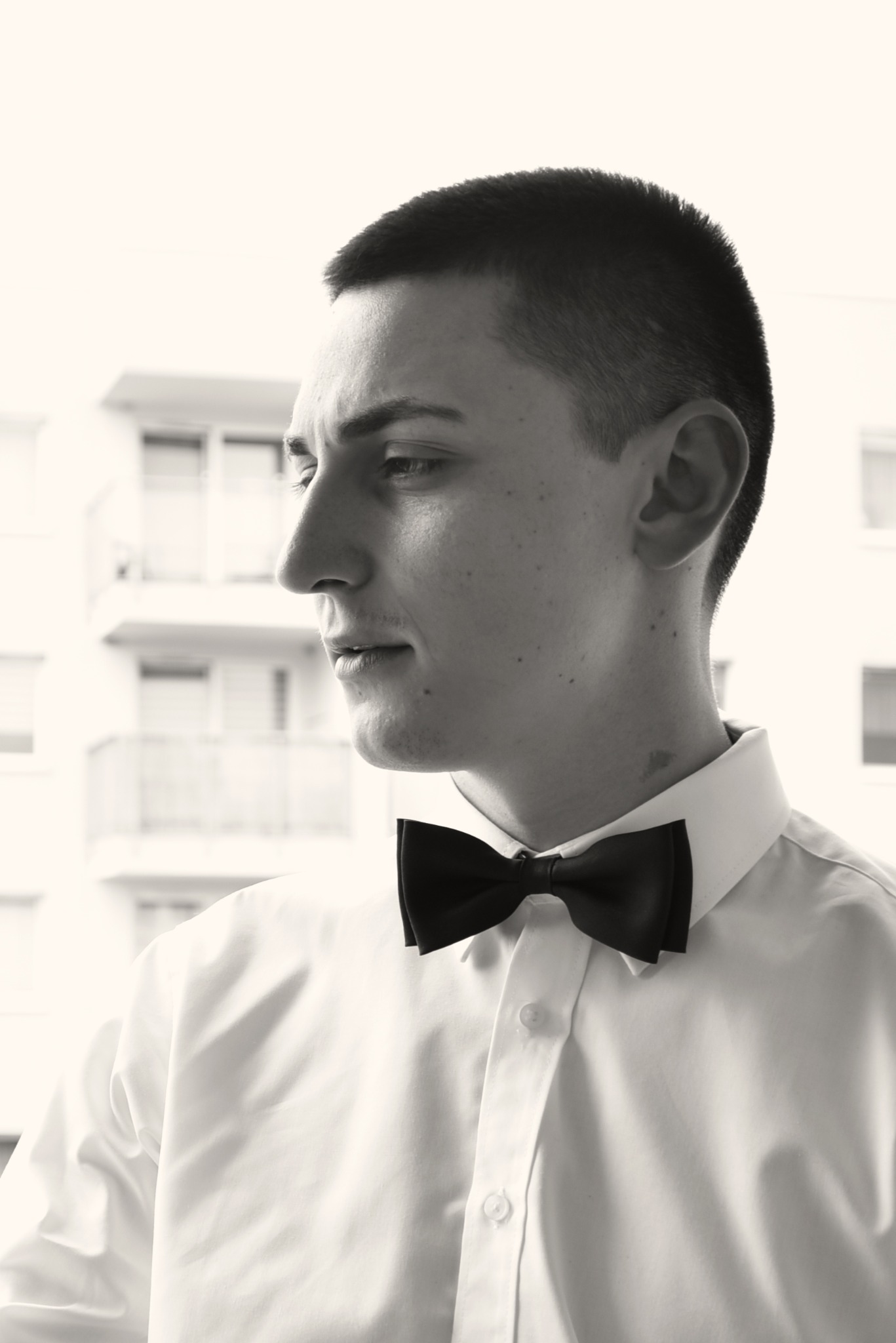 Czarno-białe, profilowe ujęcie młodego mężczyzny w białej koszuli i muszce, z budynkiem w tle. Delikatne światło podkreśla rysy twarzy.