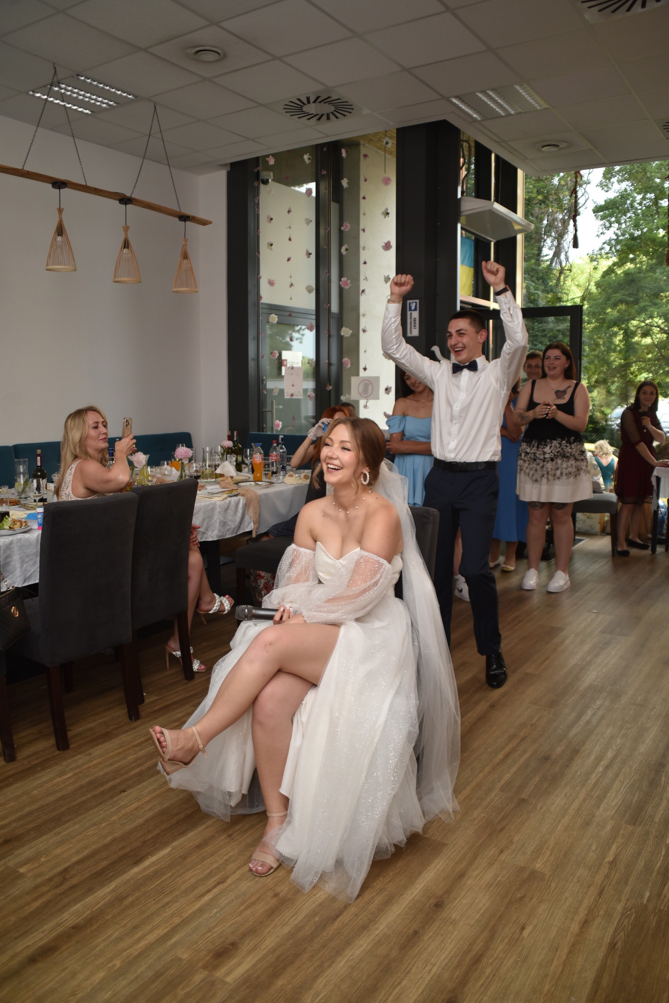 Uśmiechnięta panna młoda siedzi na krześle z mikrofonem, obok niej mężczyzna z podniesionymi rękami, w tle goście weselni i dekoracje kwiatowe.