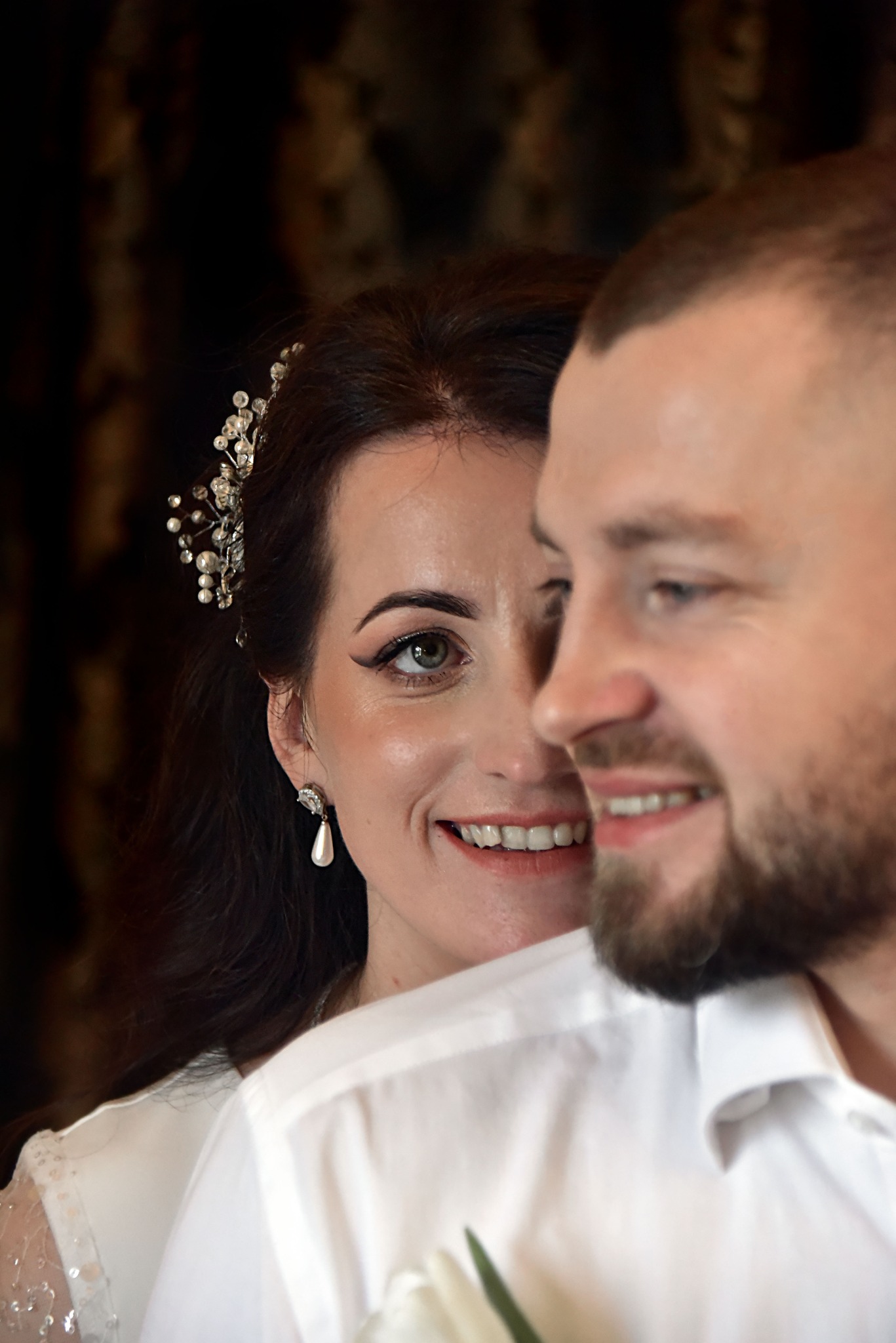 Uśmiechnięta panna młoda z ozdobą we włosach i perłowym kolczykiem, spogląda zza ramienia pana młodego w białej koszuli. Portret w ciepłych tonach.