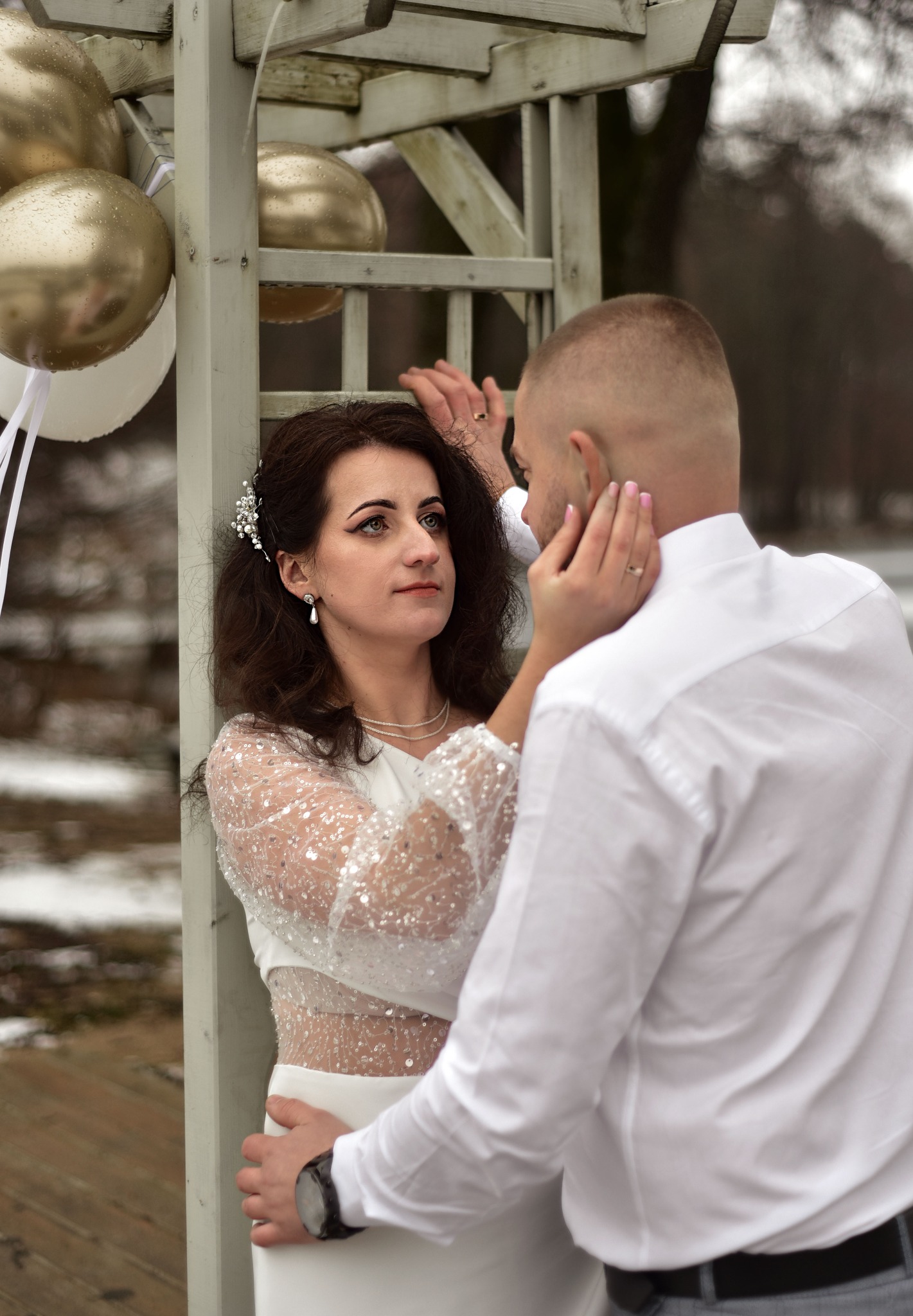 Para młoda w białych strojach pozuje przy drewnianej konstrukcji z balonami. Panna młoda z ozdobą we włosach patrzy w lewo, pan młody obejmuje ją. Zimowa sceneria.