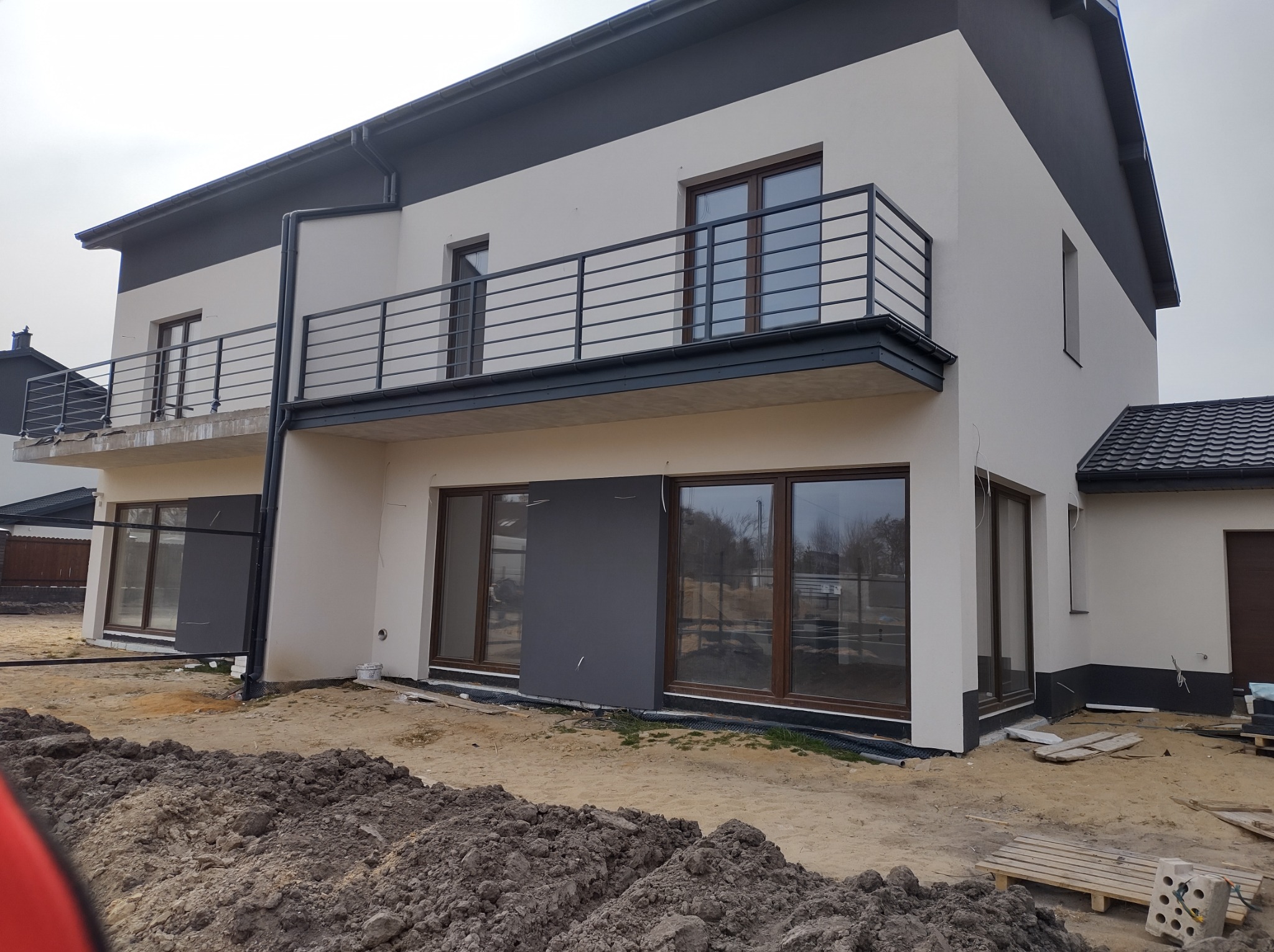 Nowoczesny dom dwukondygnacyjny z balkonami i dużymi oknami, w trakcie prac wykończeniowych. Widoczne ziemne prace wokół budynku i palety budowlane.