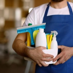 Agnieszka Olejnik - Mężczyzna w niebieskim fartuchu trzyma biały pojemnik z przyborami do sprzątania: płyny, ściągaczka, gąbki. Rozmyte tło biurowe.