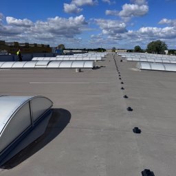 CONVION SPÓŁKA Z OGRANICZONĄ ODPOWIEDZIALNOŚCIĄ - Montaż świetlików dachowych na płaskim dachu przemysłowym. Widoczni pracownicy w odblaskowych kamizelkach. Szary dach, błękitne niebo z chmurami.
