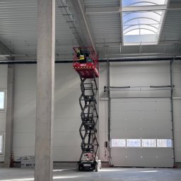 CONVION SPÓŁKA Z OGRANICZONĄ ODPOWIEDZIALNOŚCIĄ - Pracownik na podnośniku koszowym instaluje system podwieszeń pod sufitem w hali. Widoczne elementy konstrukcyjne i brama garażowa.