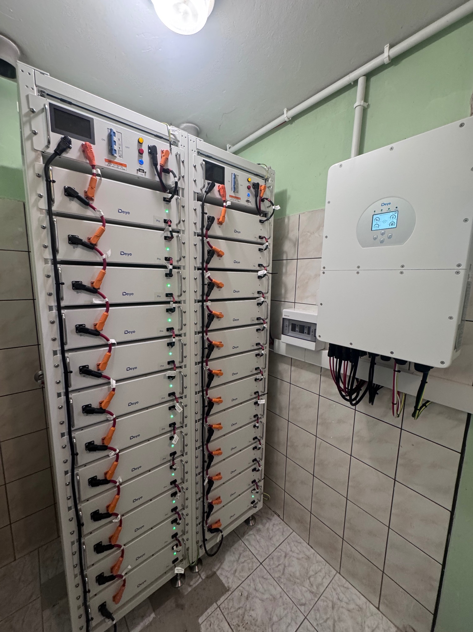 Bateria magazynu energii Deye Pro w pomieszczeniu z kafelkami, obok falownik. Widoczne kable i złącza. Instalacja wewnątrz budynku.