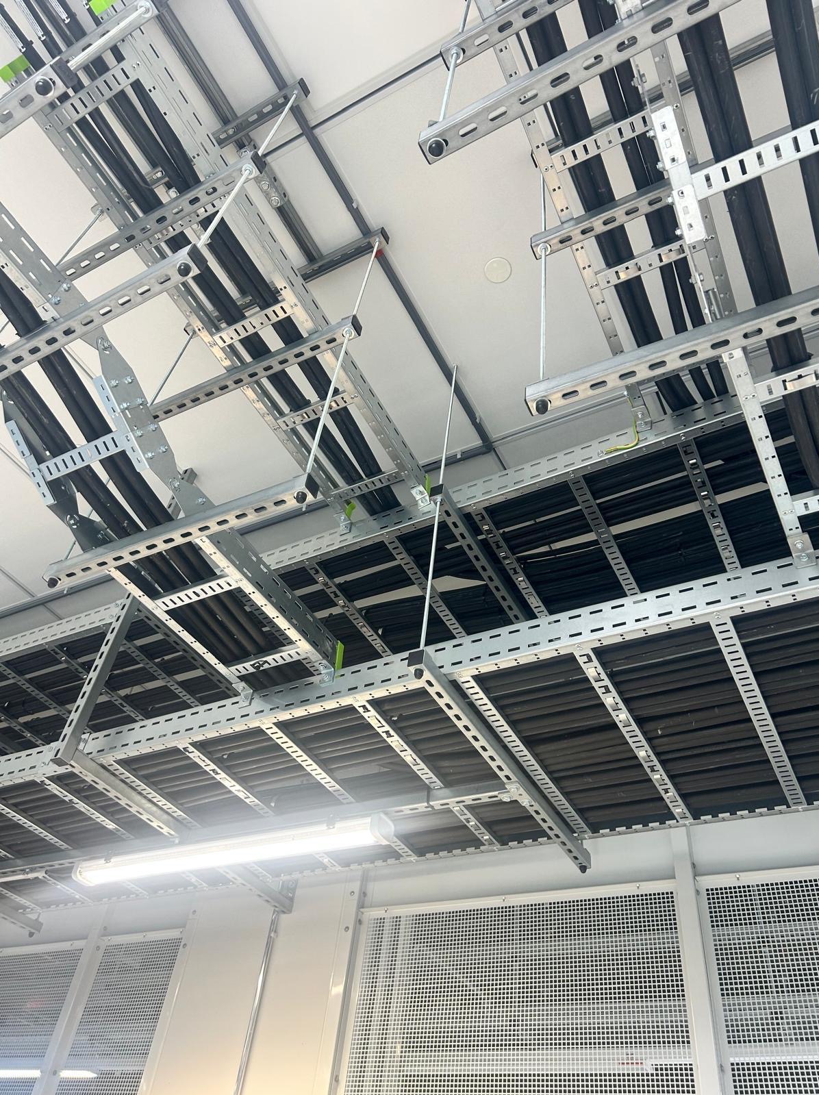 Skomplikowany system metalowych korytek kablowych pod sufitem, z widocznymi czarnymi kablami i elementami mocującymi. Oświetlenie jarzeniowe w tle.