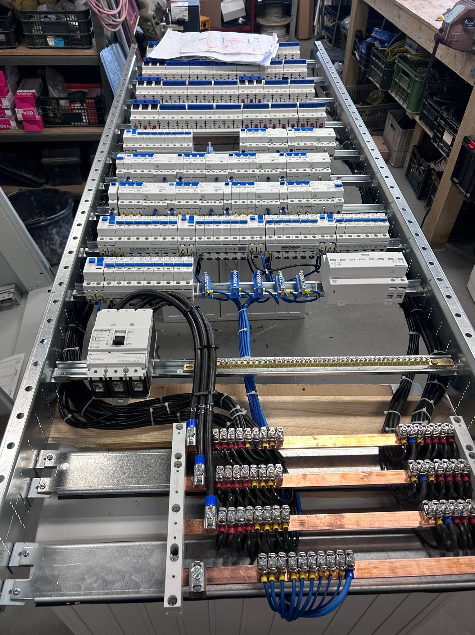 Rozdzielnica elektryczna w metalowej obudowie z licznymi zabezpieczeniami, przewodami i szynami. Widoczne elementy montażowe i schemat na górze.