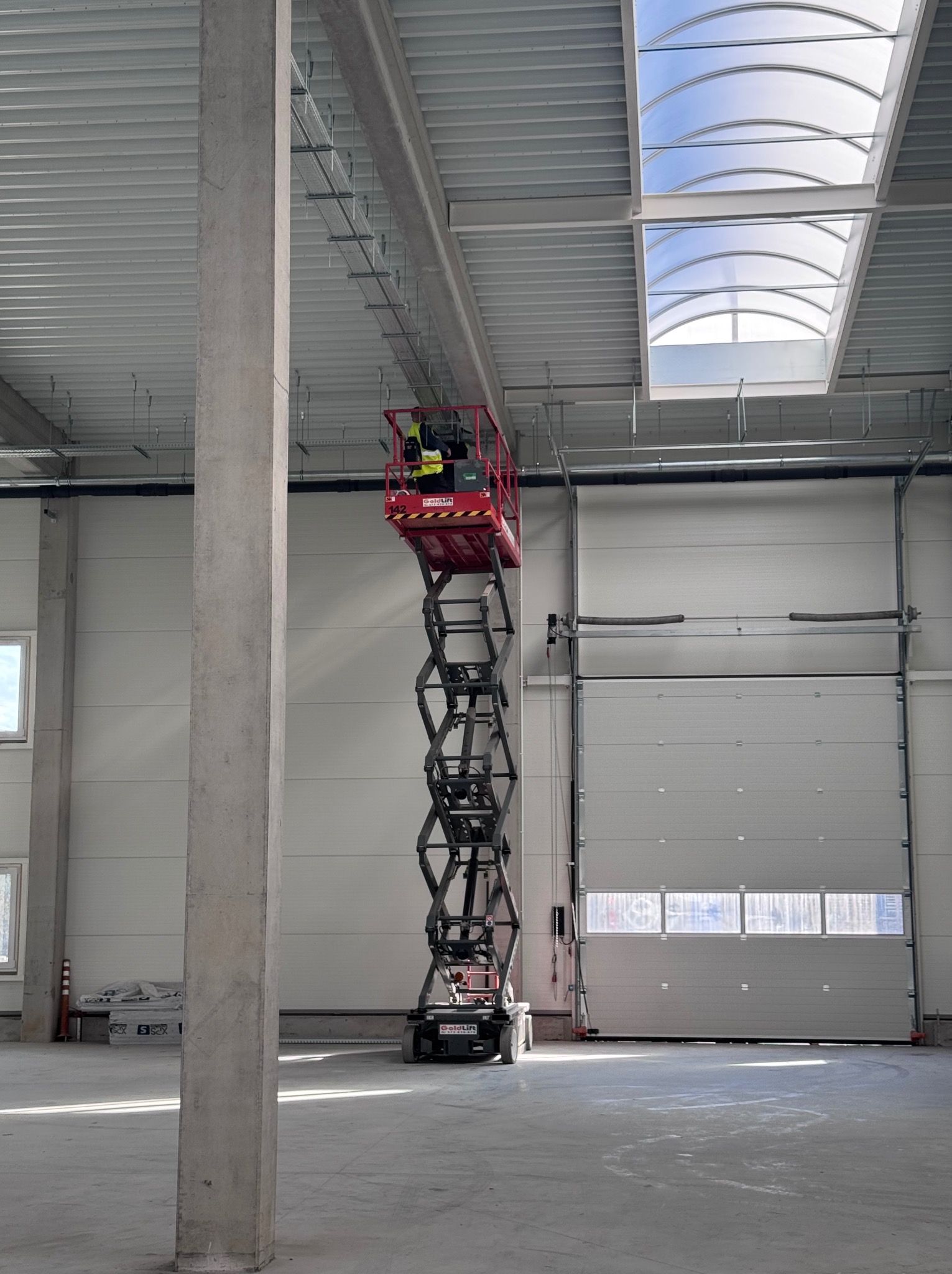 Pracownik na podnośniku koszowym instaluje system podwieszeń pod sufitem w hali. Widoczne elementy konstrukcyjne i brama garażowa.