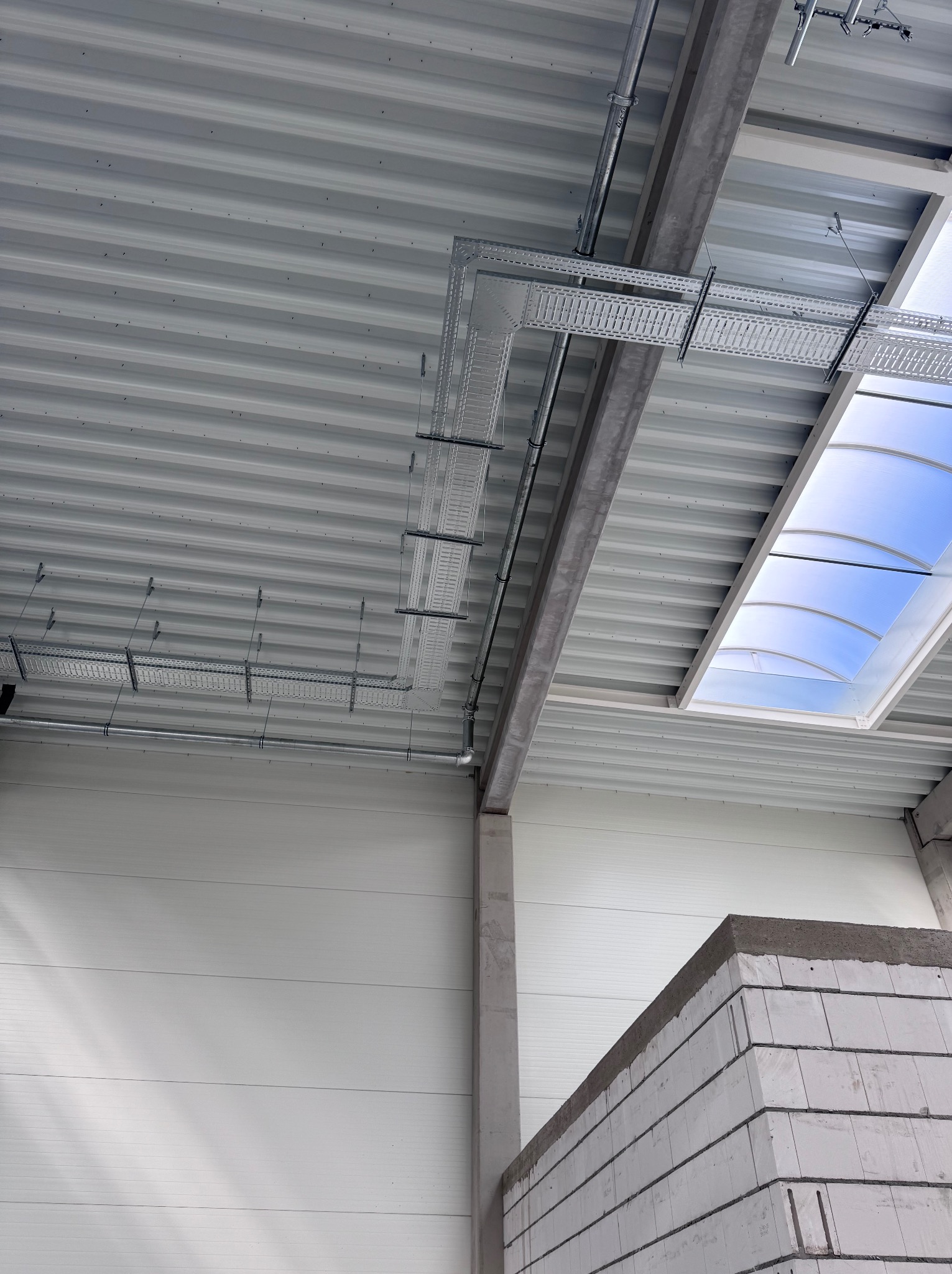 Szara, metalowa konstrukcja sufitu z zamontowanymi korytami kablowymi i świetlikiem w hali przemysłowej, widok z dołu, fragment ściany z betonu komórkowego.