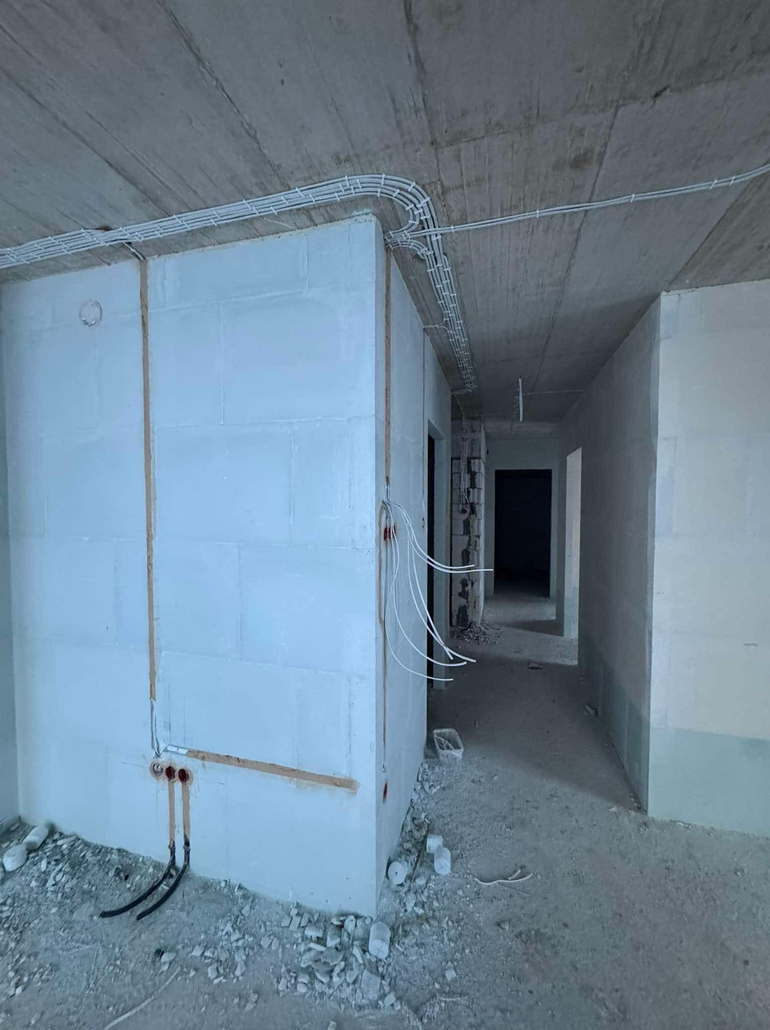Wnętrze w stanie surowym z widoczną instalacją elektryczną: kable w peszlach prowadzone po suficie i ścianach, puszki elektryczne w ścianie. Widok na korytarz.