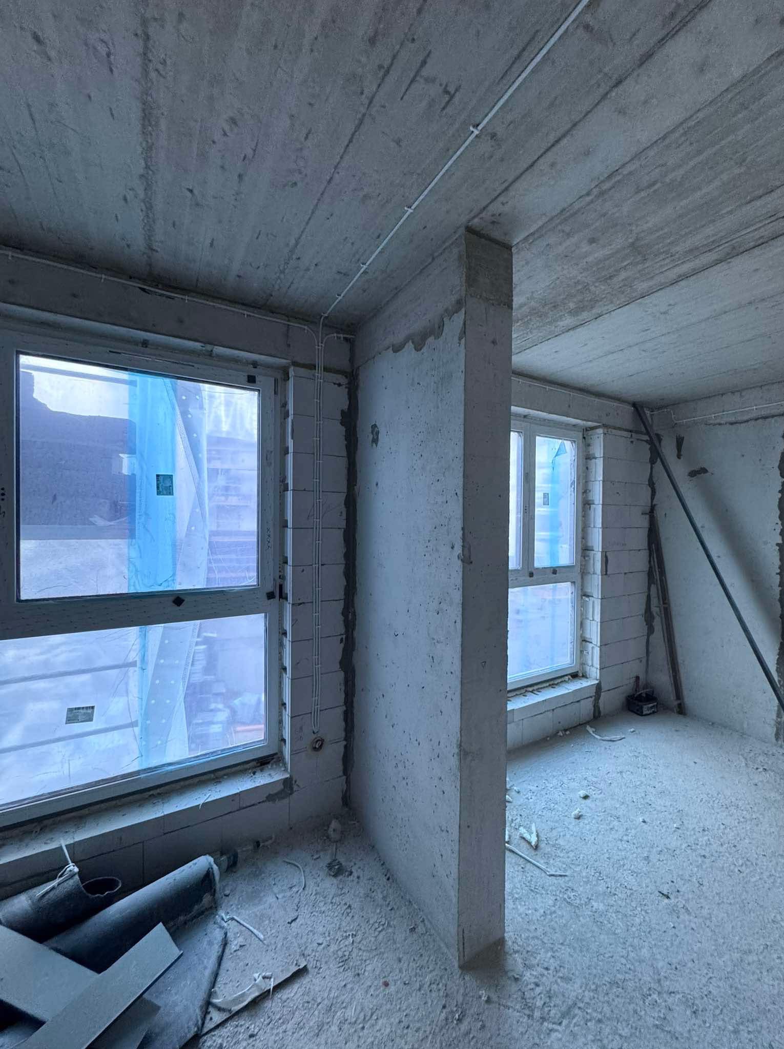 Surowy stan wnętrza: widok na dwa okna z niebieską folią ochronną, ściany z betonu komórkowego, widoczne przewody elektryczne, podłoga pokryta pyłem budowlanym.