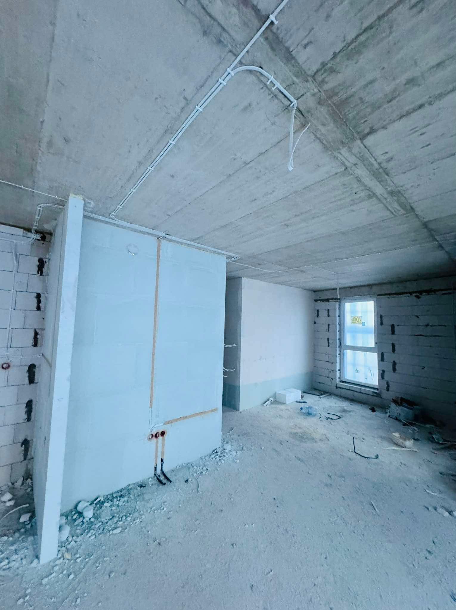 Surowy stan wnętrza: widoczne przewody elektryczne pod sufitem, ściany z bloczków i okno. Wnętrze w trakcie prac wykończeniowych, z widocznymi elementami instalacji.