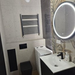 Lucas - Nowoczesna łazienka z czarną armaturą, umywalką na szafce, podświetlanym lustrem i grzejnikiem drabinkowym na tle płytek w odcieniach szarości i beżu.